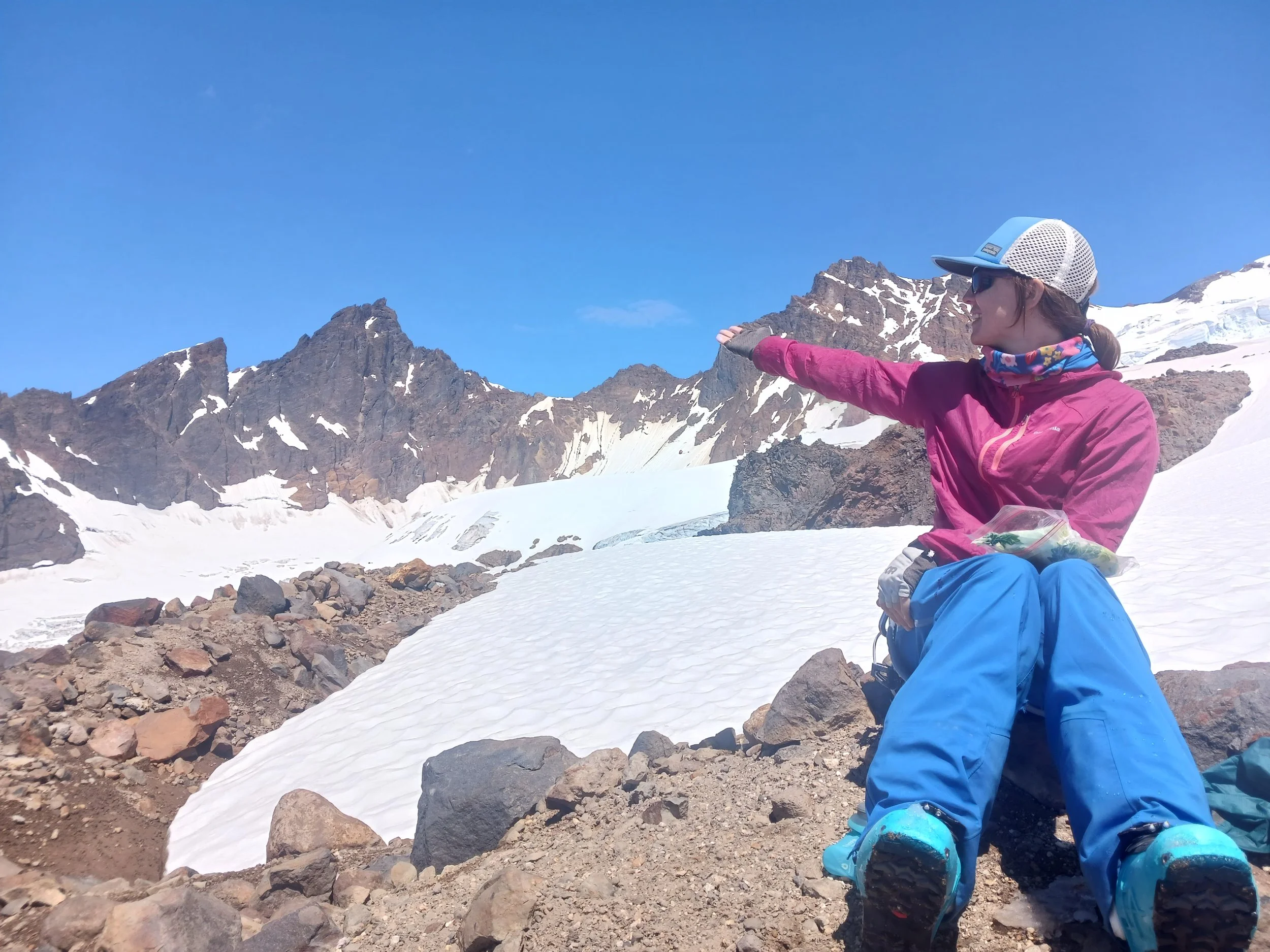 A woman wearing outdoor gear, including a pink jacket, blue pants, and a white cap, sits on rocky terrain in a snowy mountain landscape under a clear blue sky, stretching her arm forward.