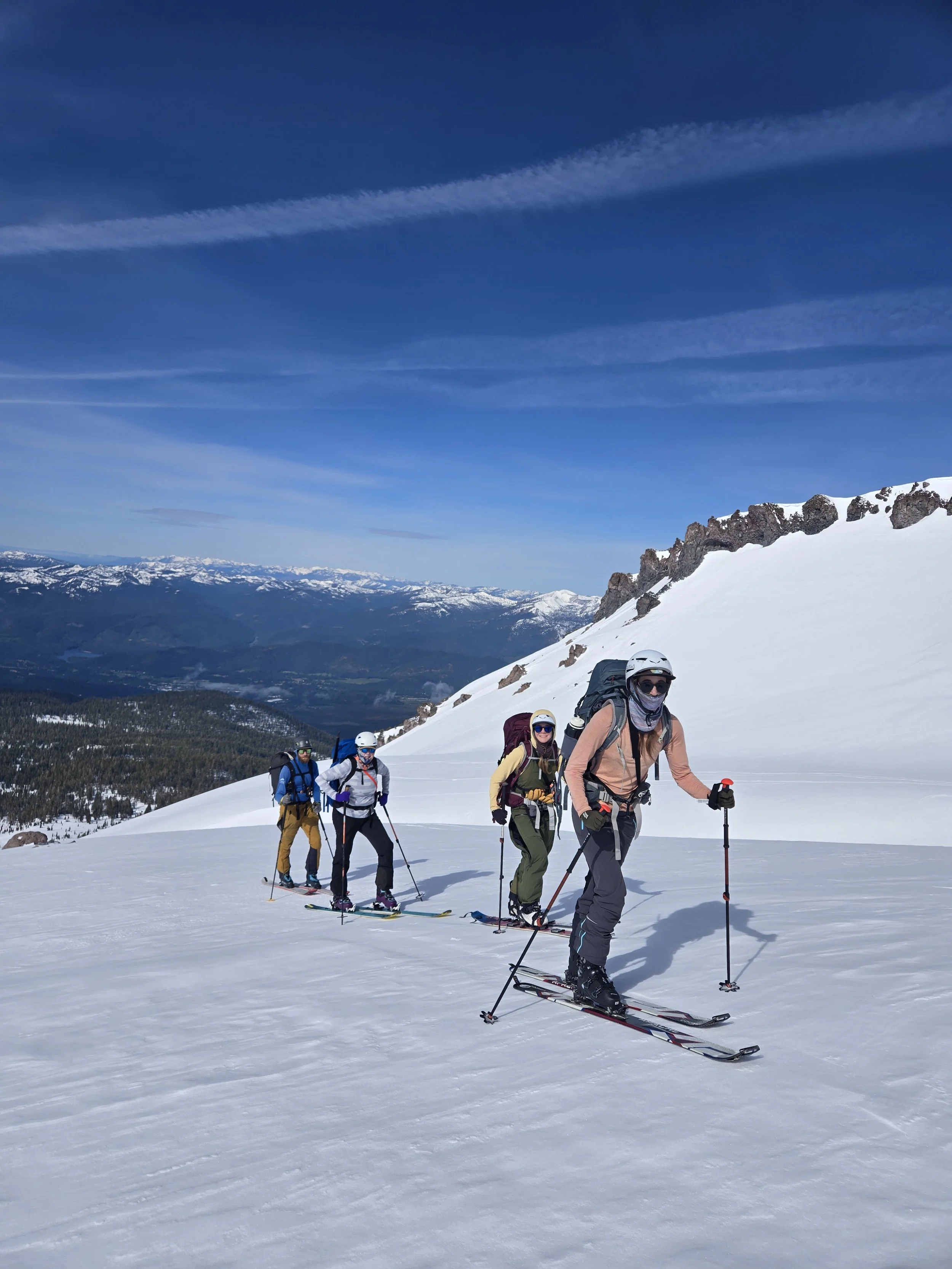 Group of four people in snow jackets and helmets skiing uphill on a snowy mountain with a clear blue sky and distant snow-capped mountains.