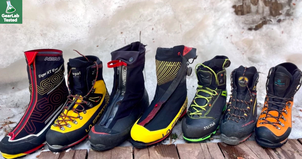 A row of six different outdoor hiking and mountaineering boots placed on a wooden surface with snow in the background.