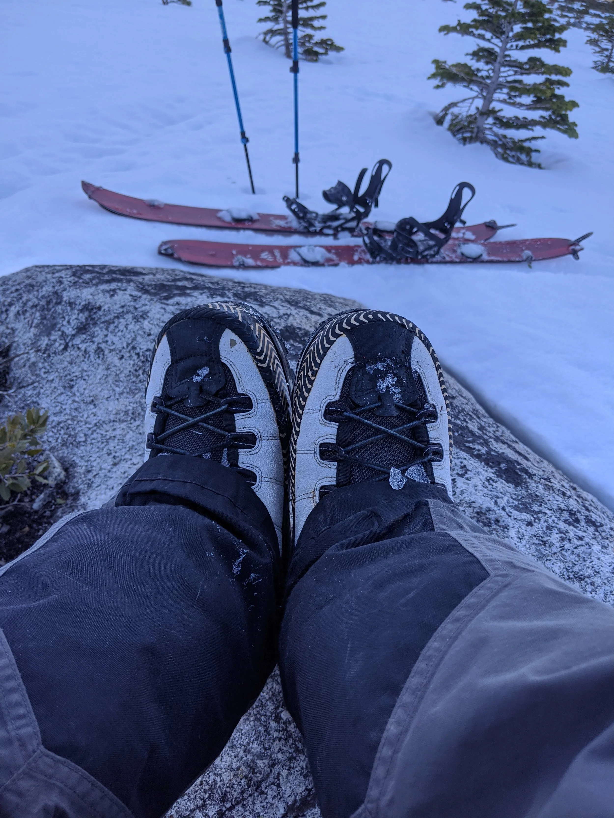 Person wearing hiking boots sitting on a rock in the snow, with a pair of red skis and ski poles lying in the snow nearby.