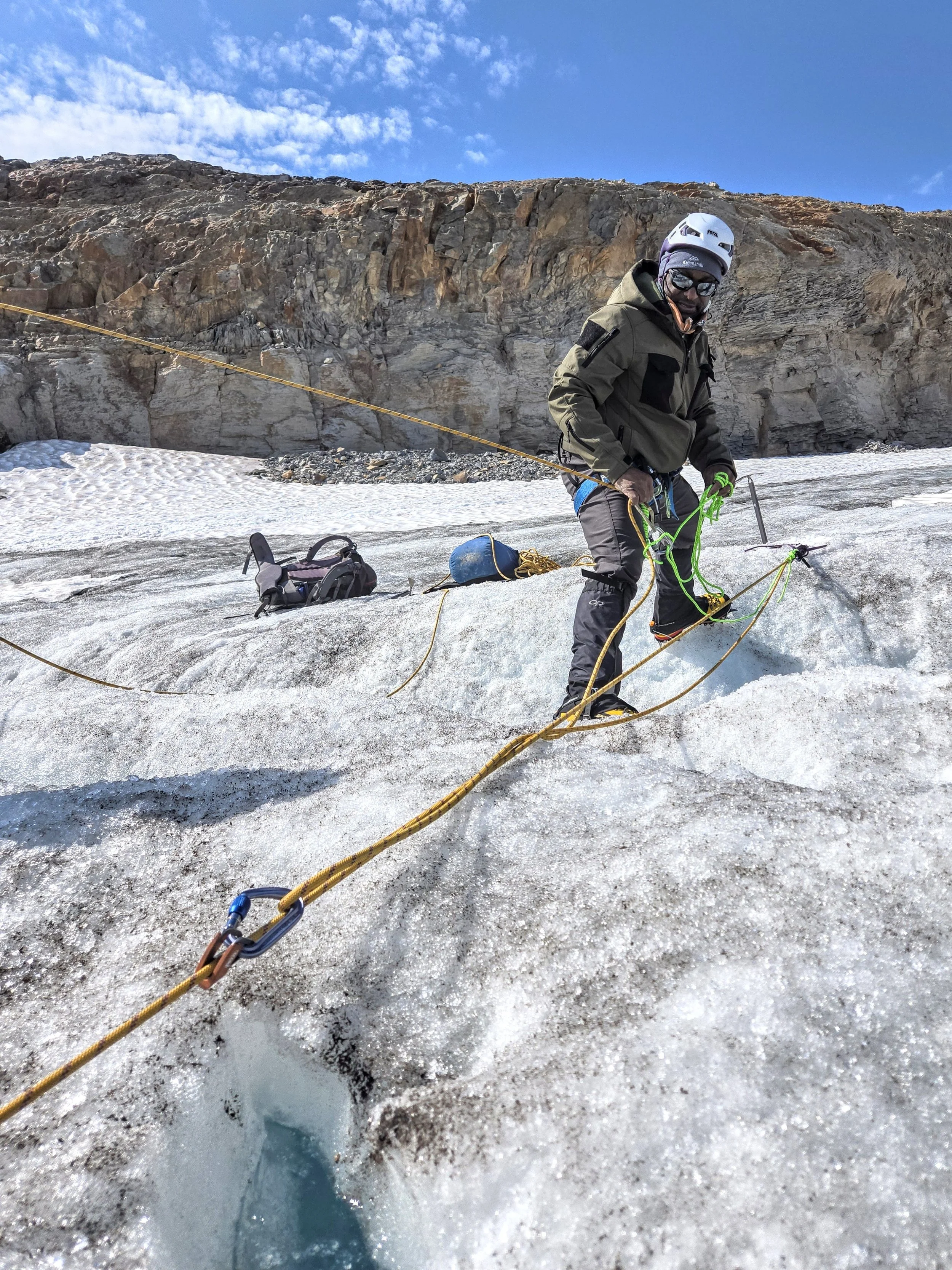 A climber in warm clothing, helmet, and goggles is securing ropes on a glacier, with a backpack and other gear nearby, against a backdrop of rocky mountains and a partly cloudy sky.