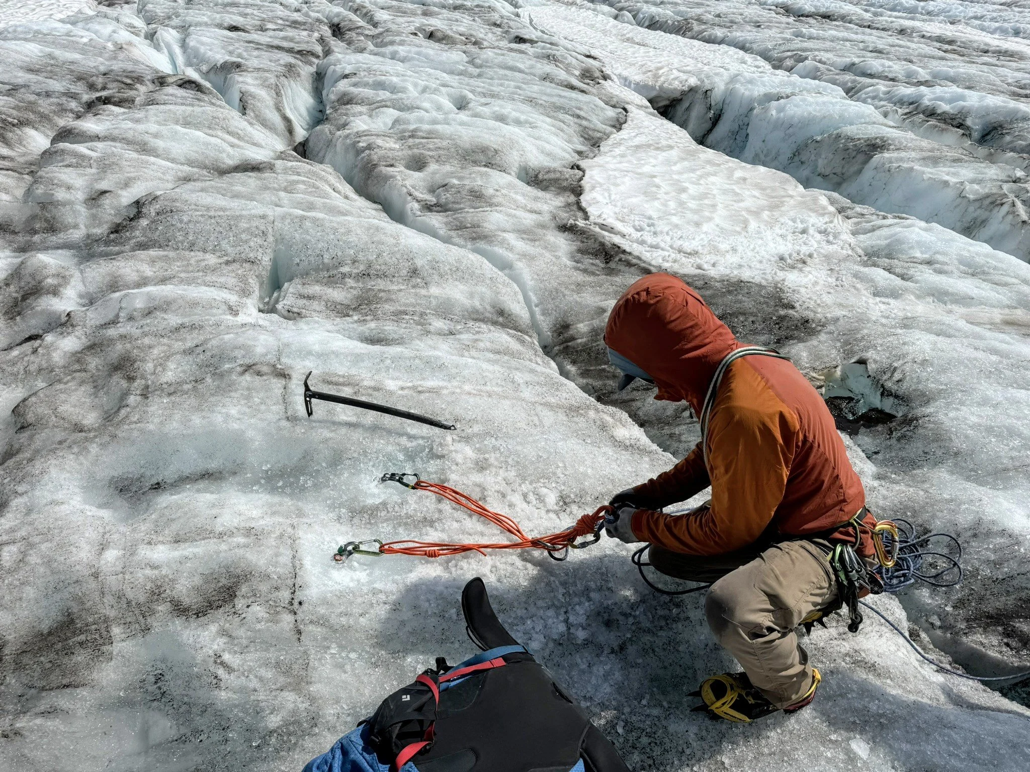 A person in orange and brown clothing, wearing a red hood, is securing climbing gear on icy terrain, with a glacier in the background, using ice axes and ropes.