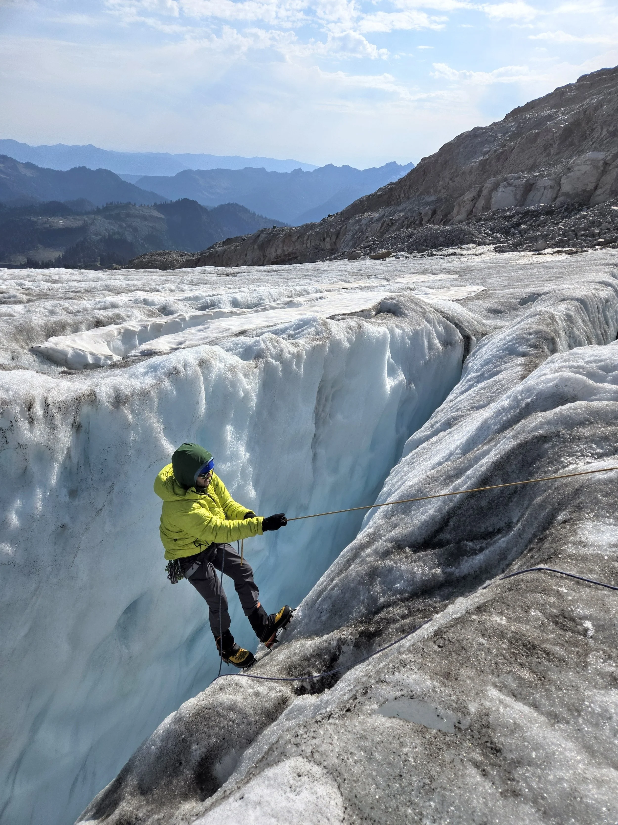 A person dressed in yellow and gray, wearing a green hood and sunglasses, is rappelling down a glacier with mountains and a blue sky in the background.