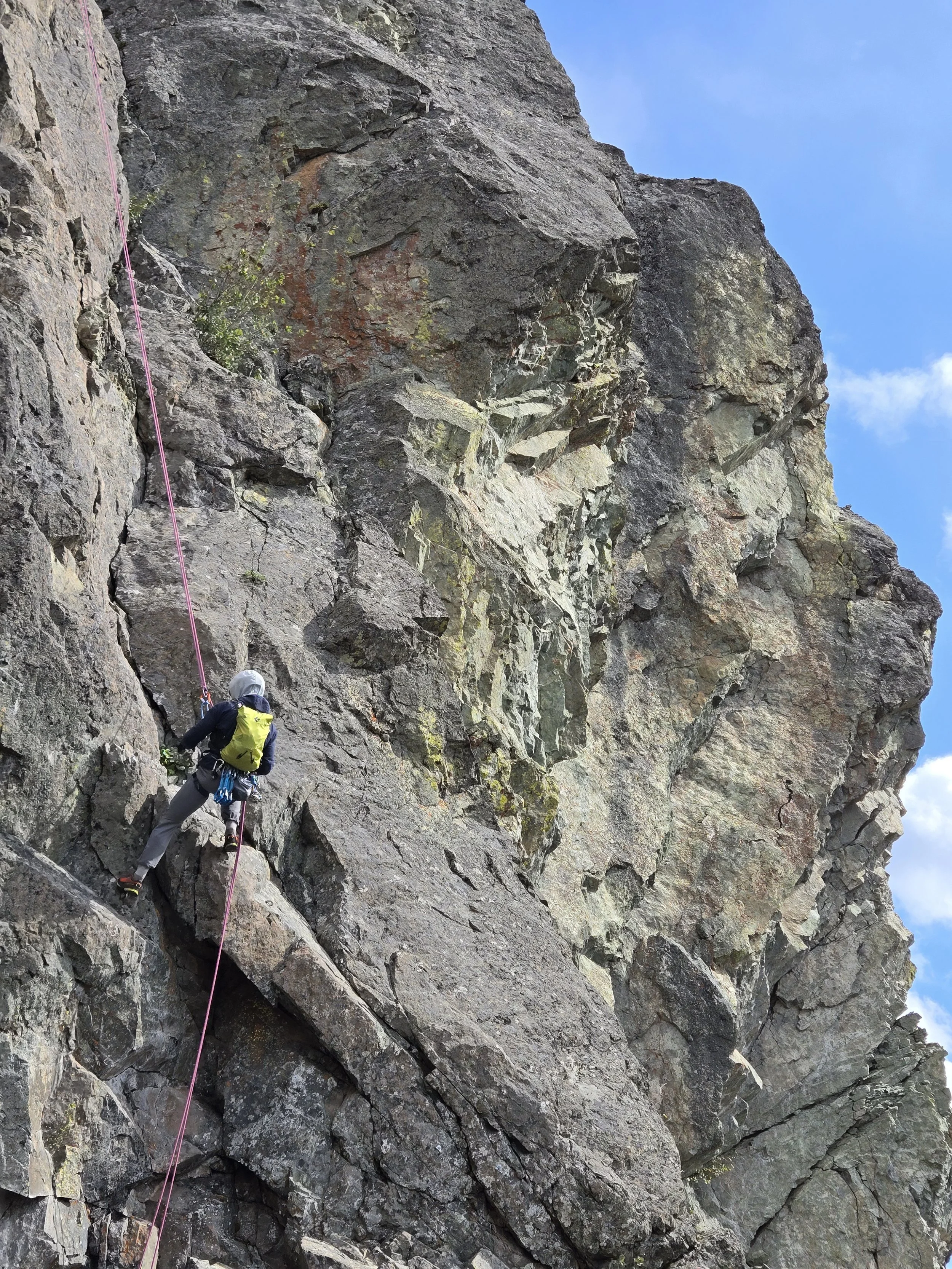 A person rock climbing on a steep mountainside with a climbing rope, harness, and gear, under a blue sky with some clouds.