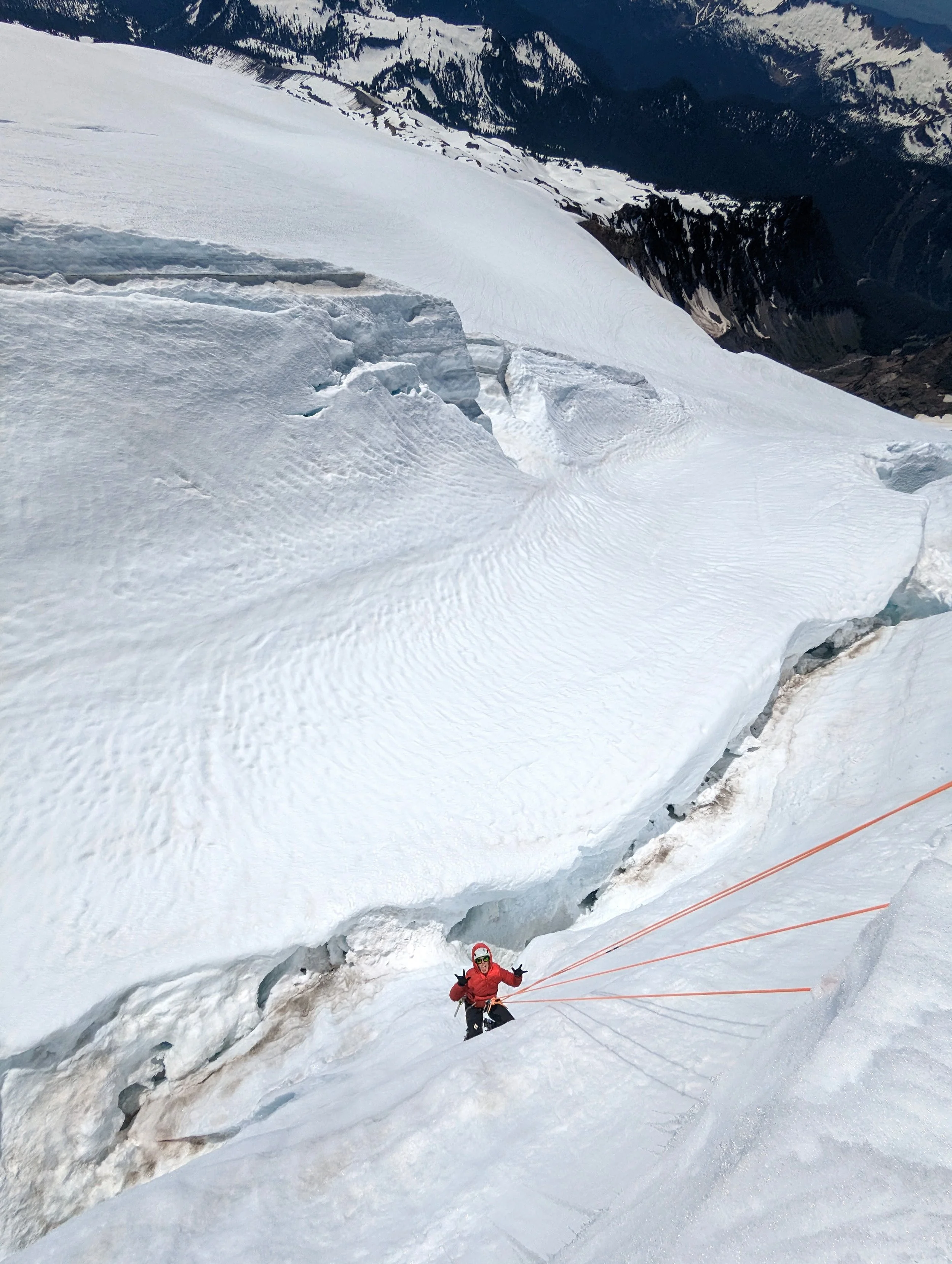 A person in red outdoor gear and a helmet is climbing a steep icy slope on a glacier with orange ropes for safety, in a mountainous snowy region.