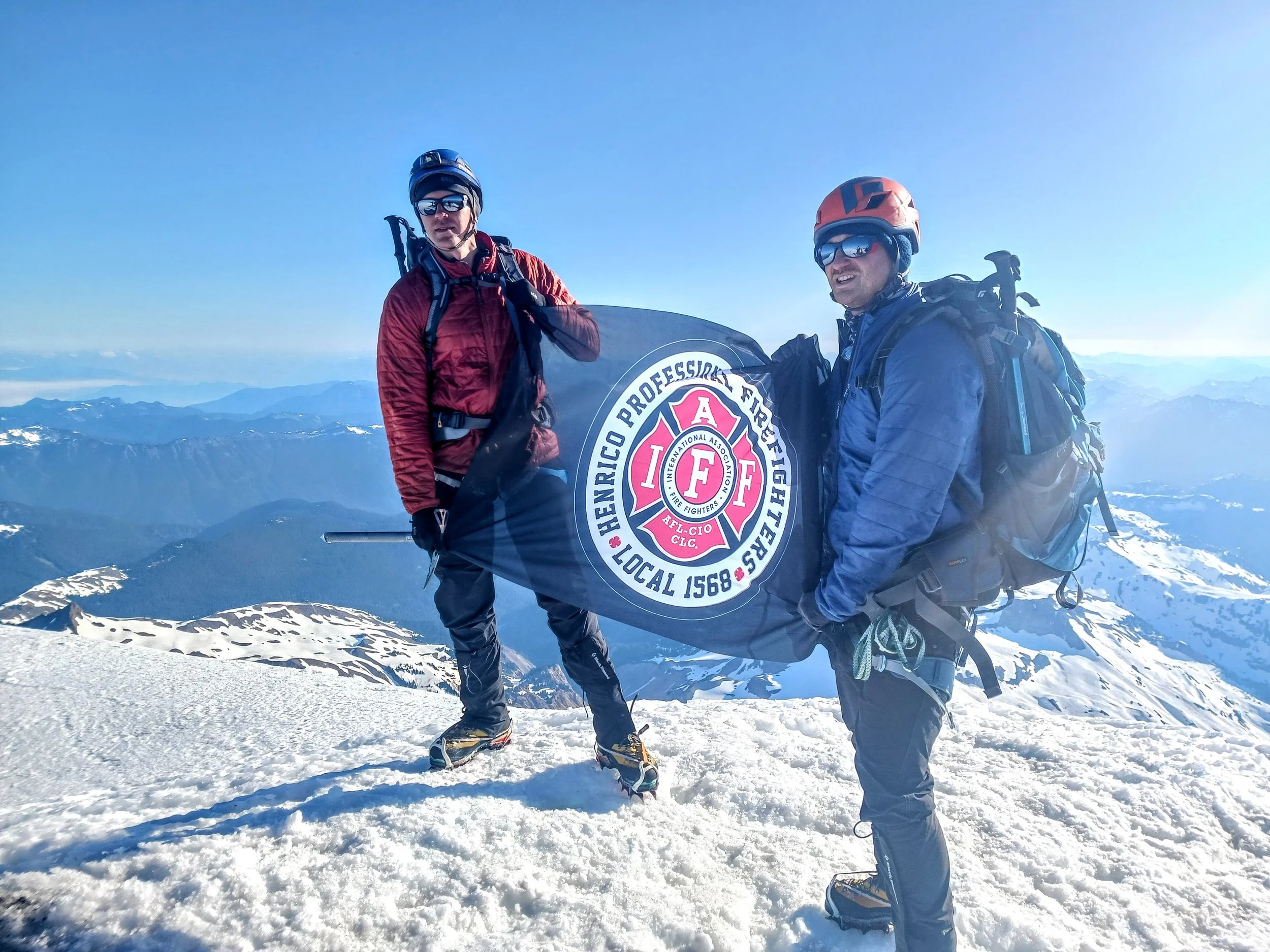 Two mountain climbers standing on snow-covered peak holding a flag with firefighter insignia, mountains in background, clear blue sky.