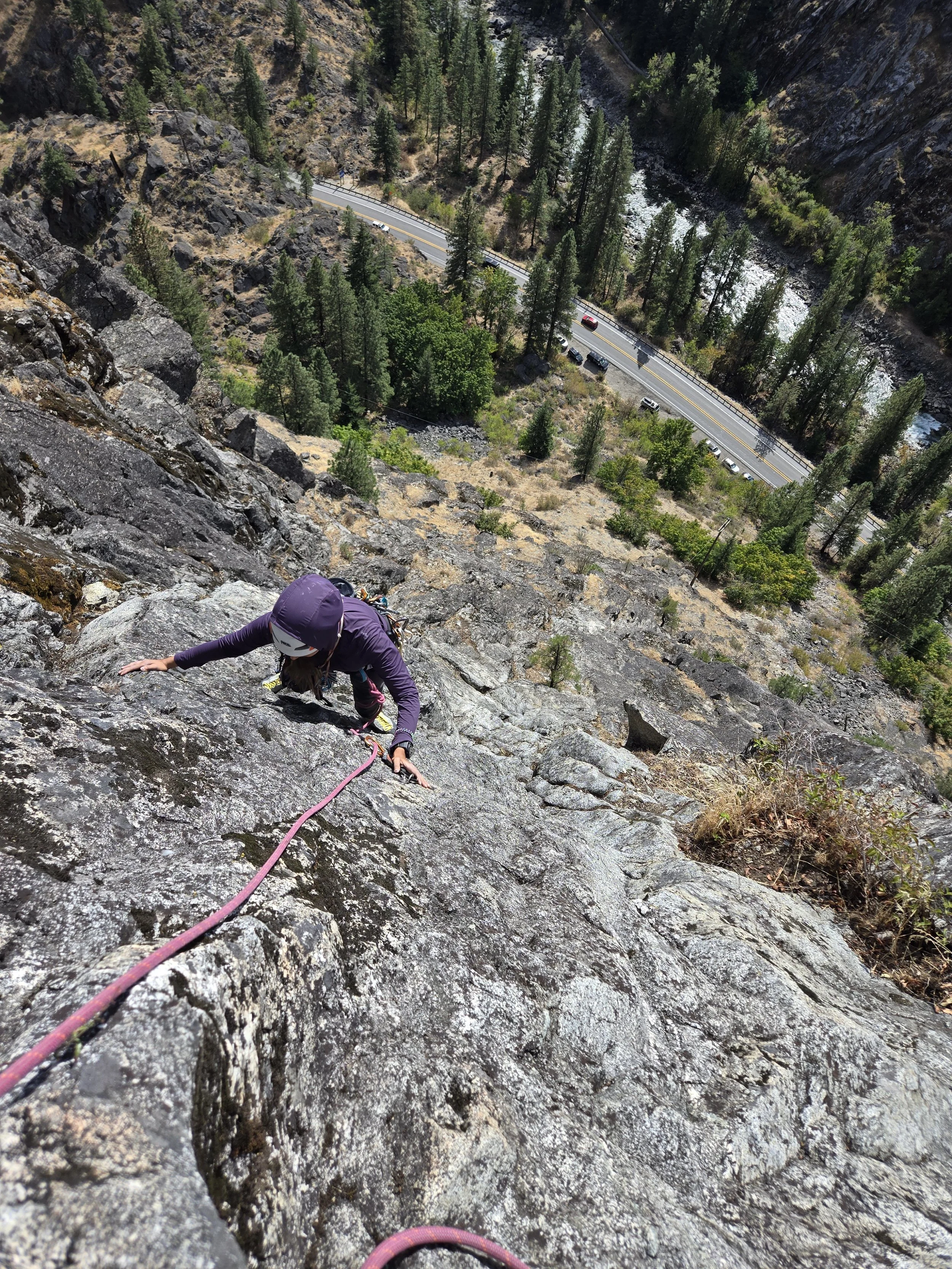 A person rock climbing on a steep cliffside near a forested valley with a river, and a road with cars at the bottom of the canyon.
