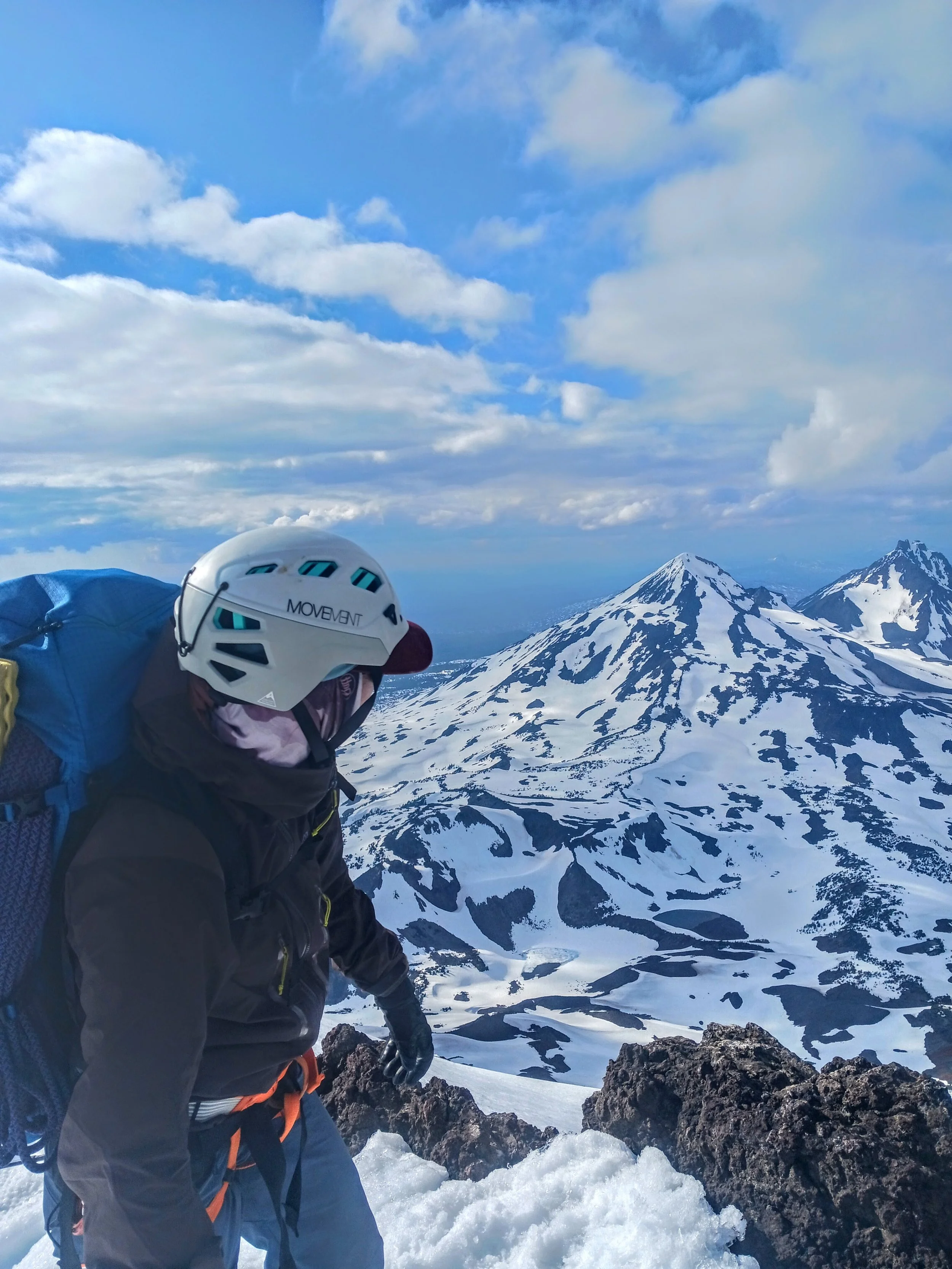 A person wearing a helmet and outdoor gear climbing snowy mountains with volcanic peaks in the background.