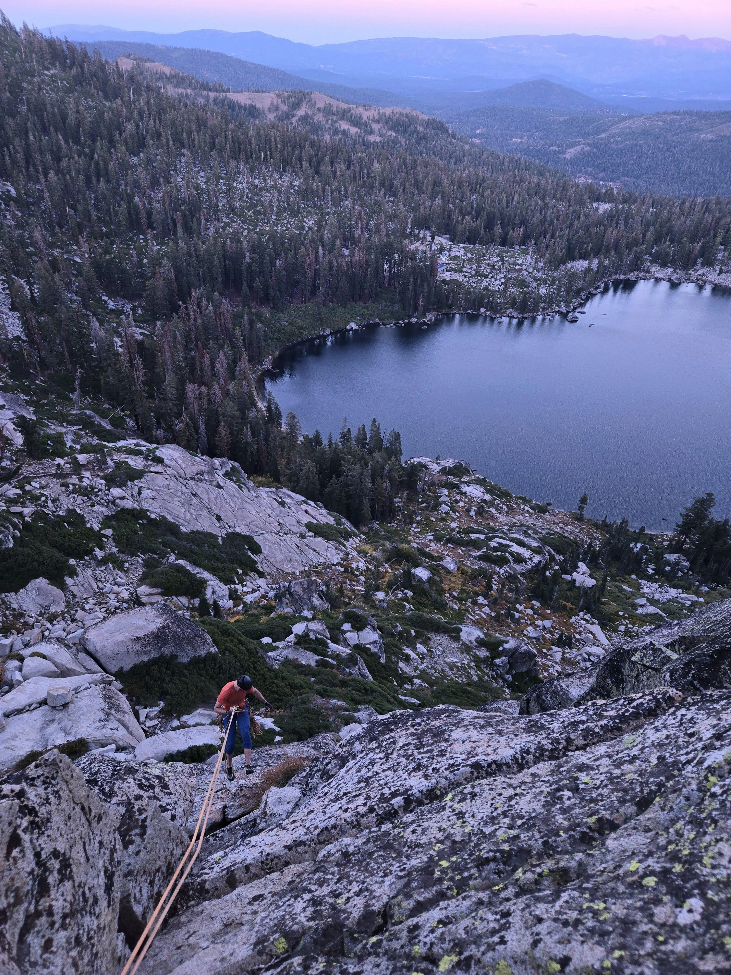 A person climbing a rocky mountain slope with a rope, overlooking a forested landscape and a lake at sunset or dusk.