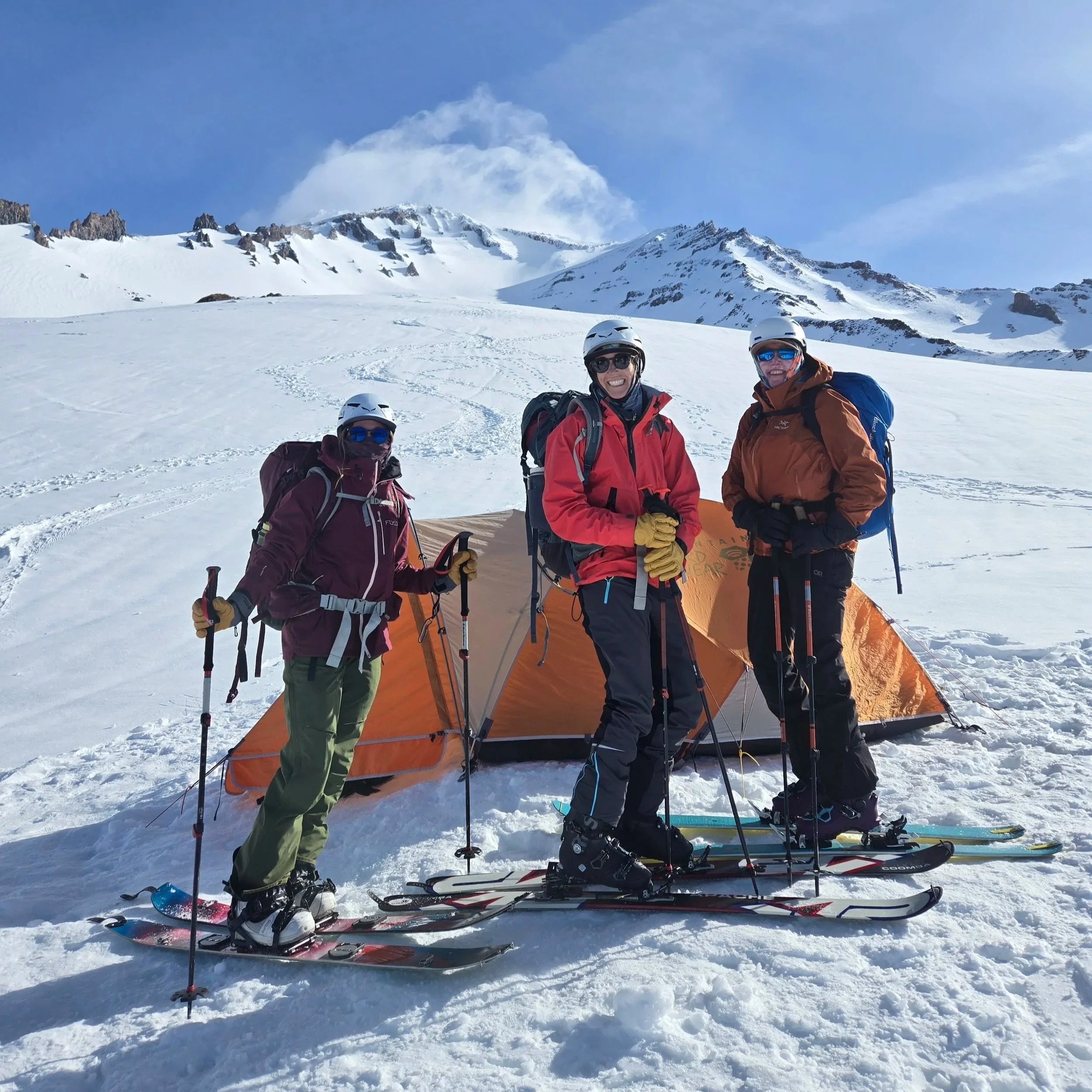 Three people in winter gear standing on snow with skis, in front of an orange tent, mountain landscape in the background.