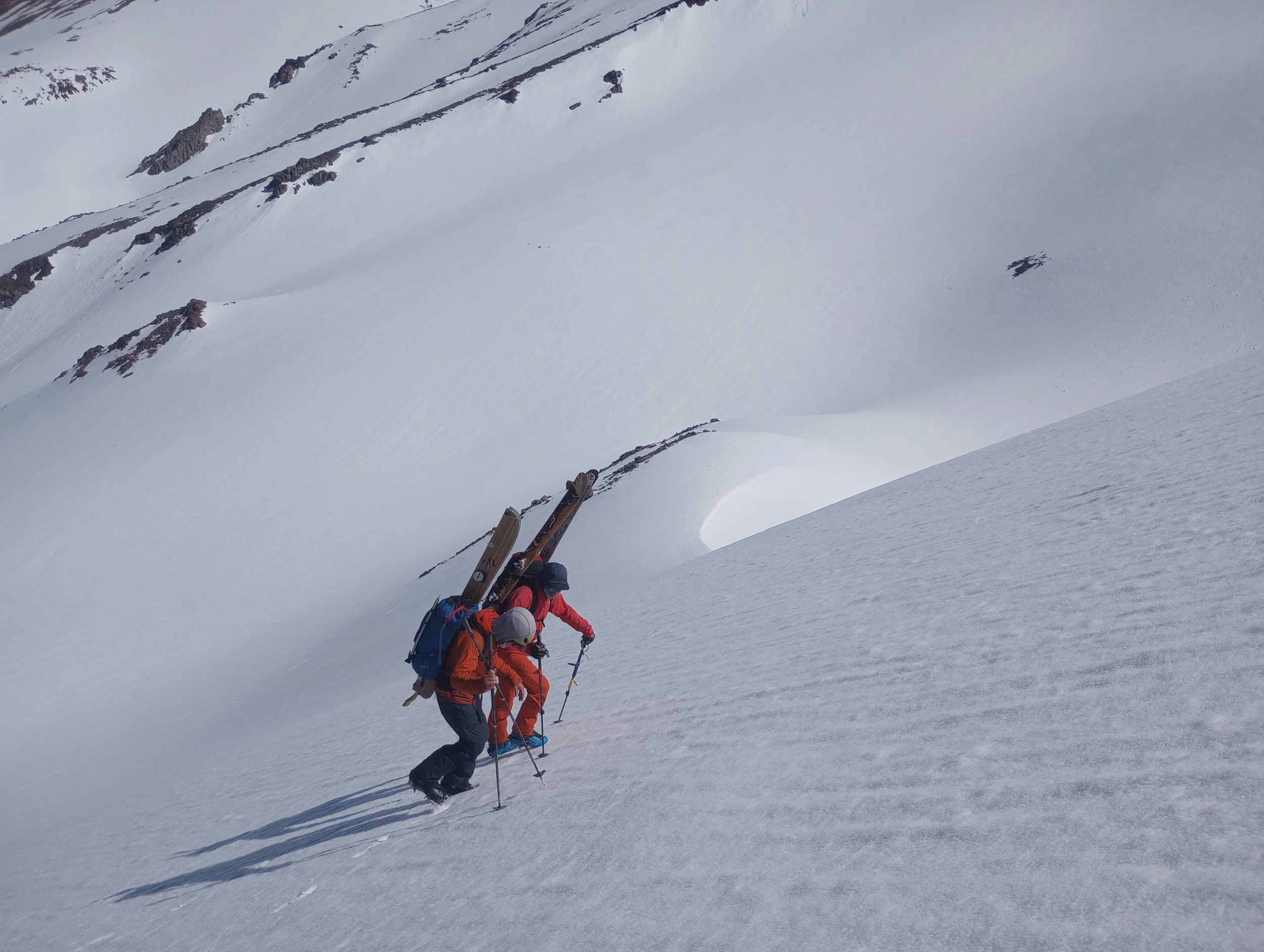 Two climbers ascending a snow-covered mountain slope with climbing gear and walking poles.