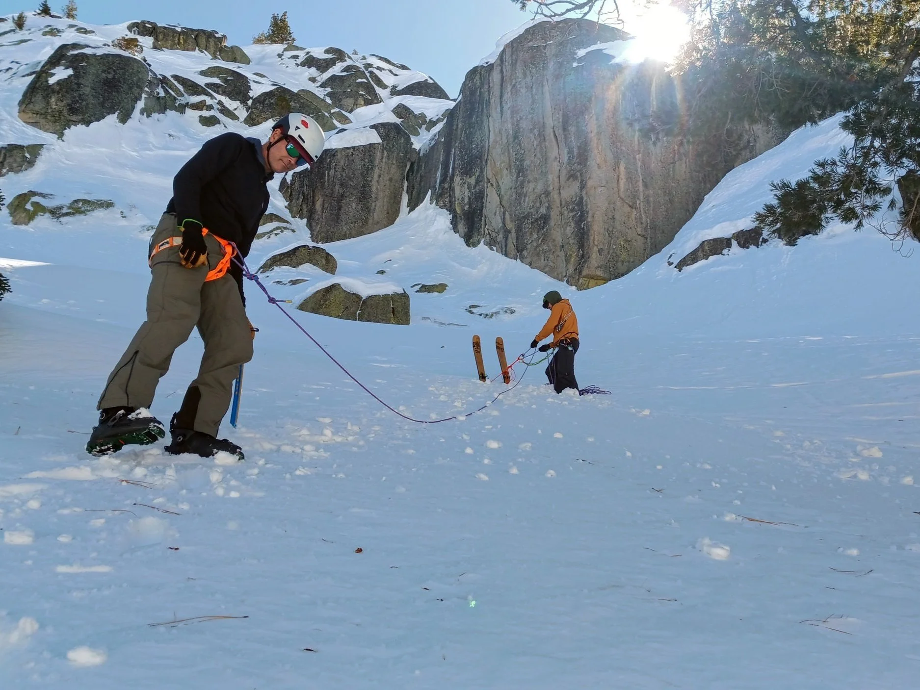 Two people in snow gear holding a rescue rope in a snowy mountain landscape with large rocks and a sunlit sky.