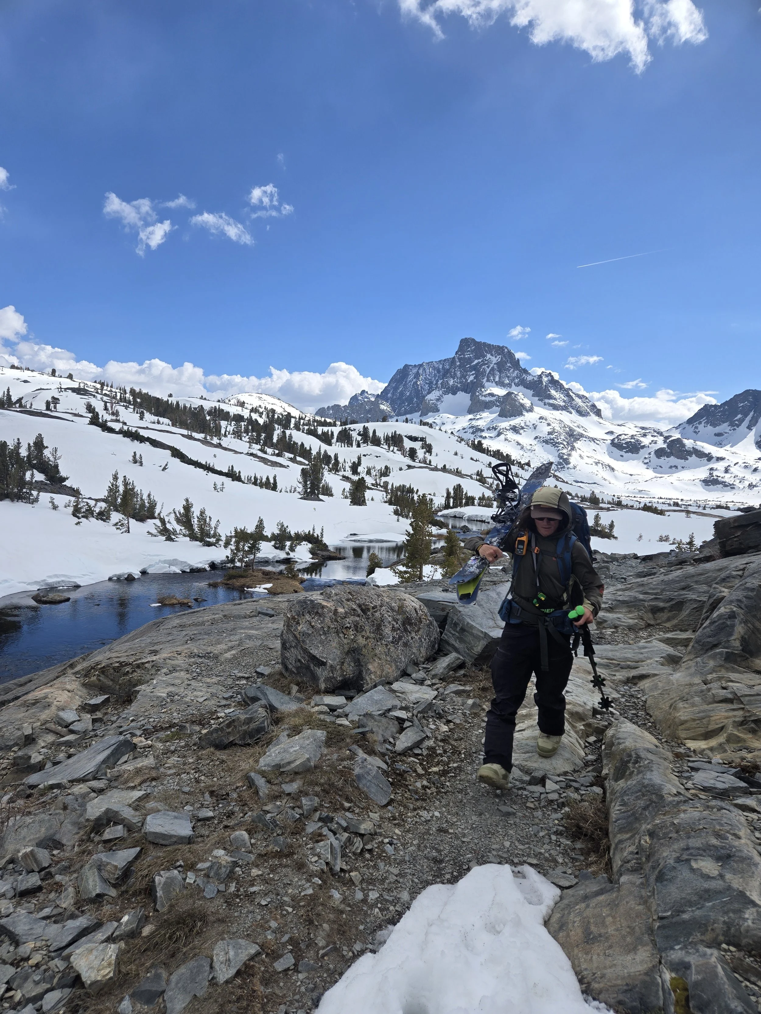 A hiker walking along a rocky trail in a snowy mountainous landscape, with a river and snow-covered trees surrounding him, under a partly cloudy blue sky.
