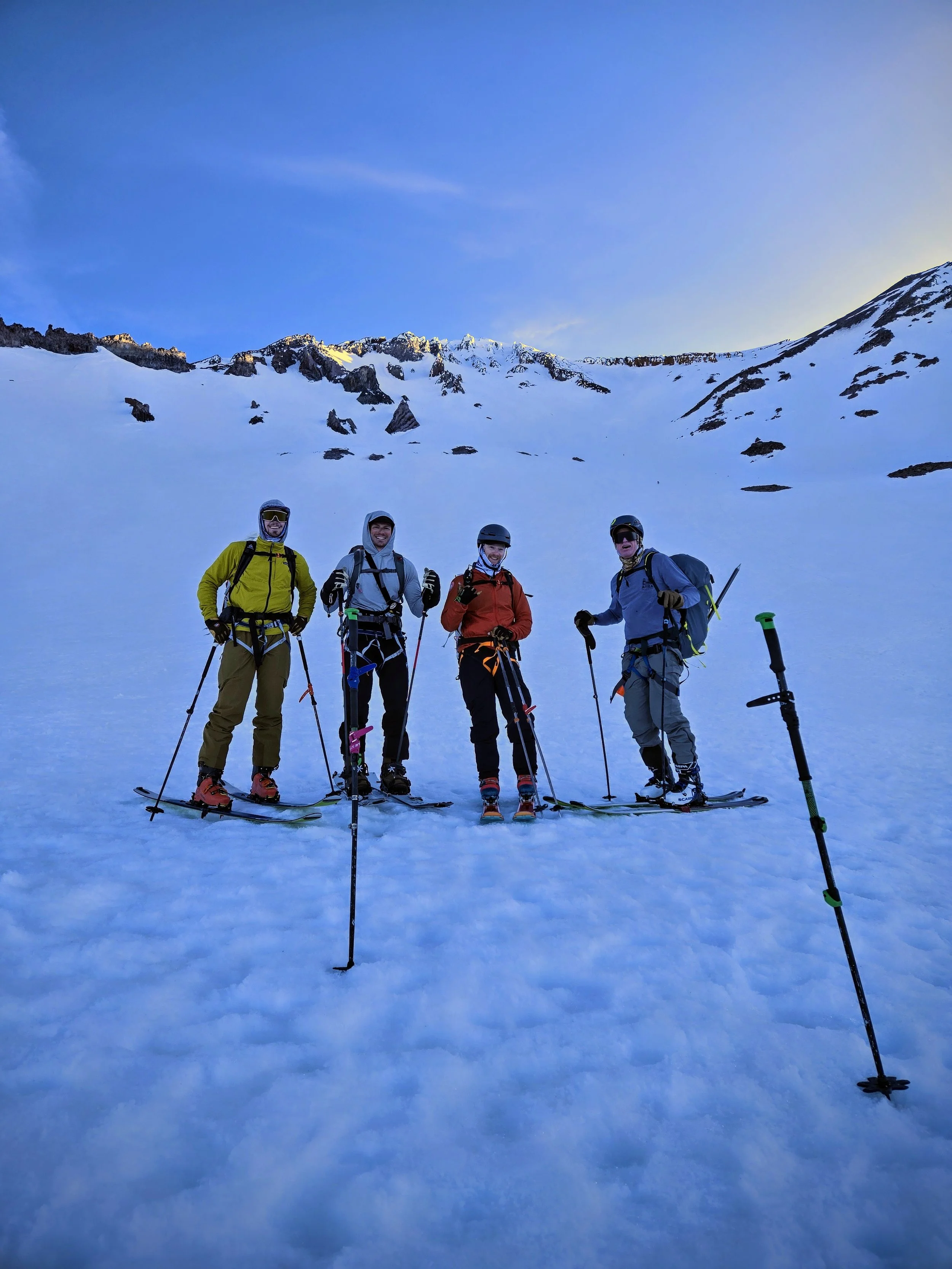 Four people dressed in winter gear, standing in snow on a mountain slope with snow-covered peaks and a clear blue sky in the background, taking a break from skiing.