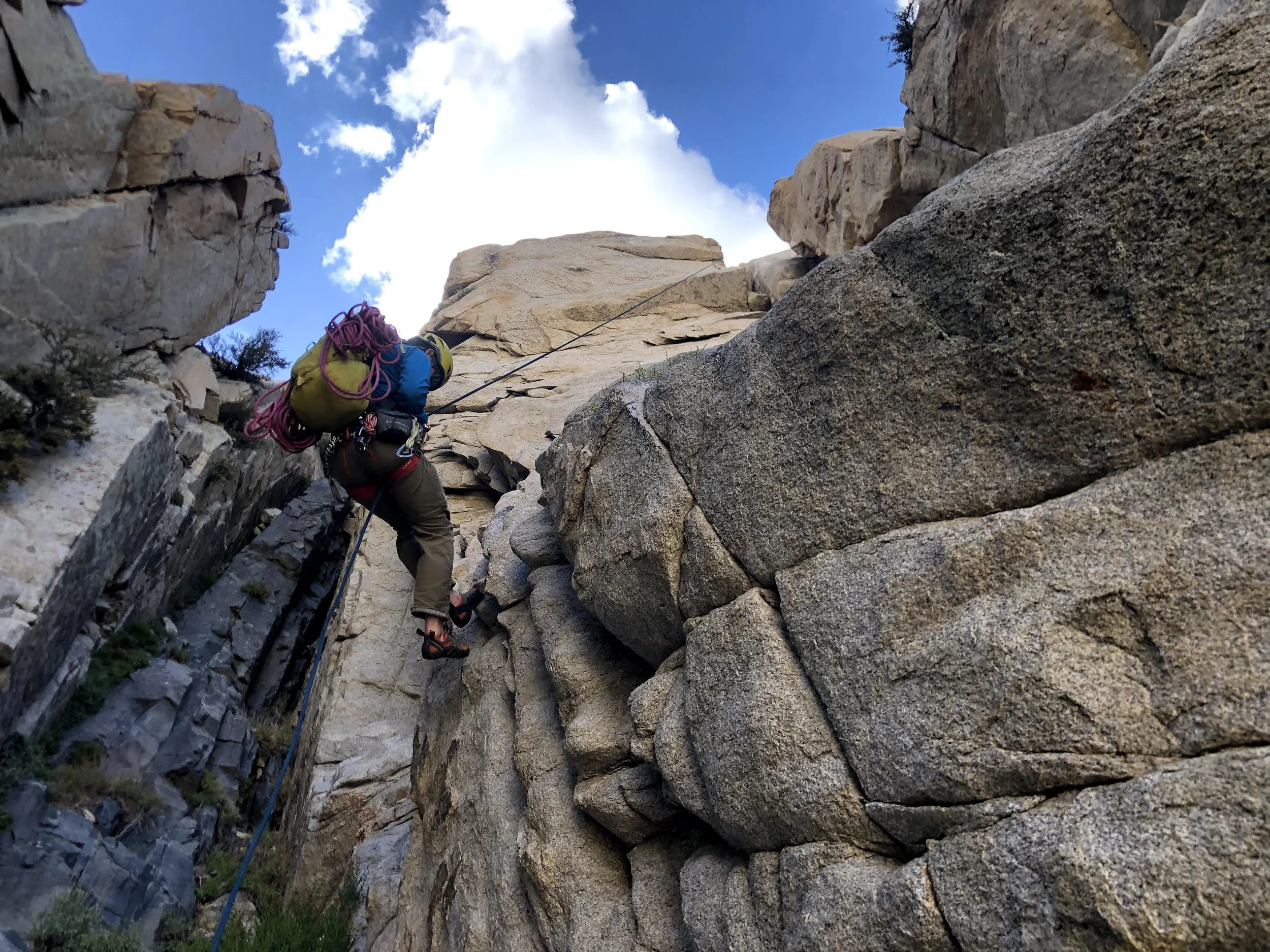 Climber ascending a rugged rock face with safety gear and a large backpack on a mountain with a partly cloudy sky.