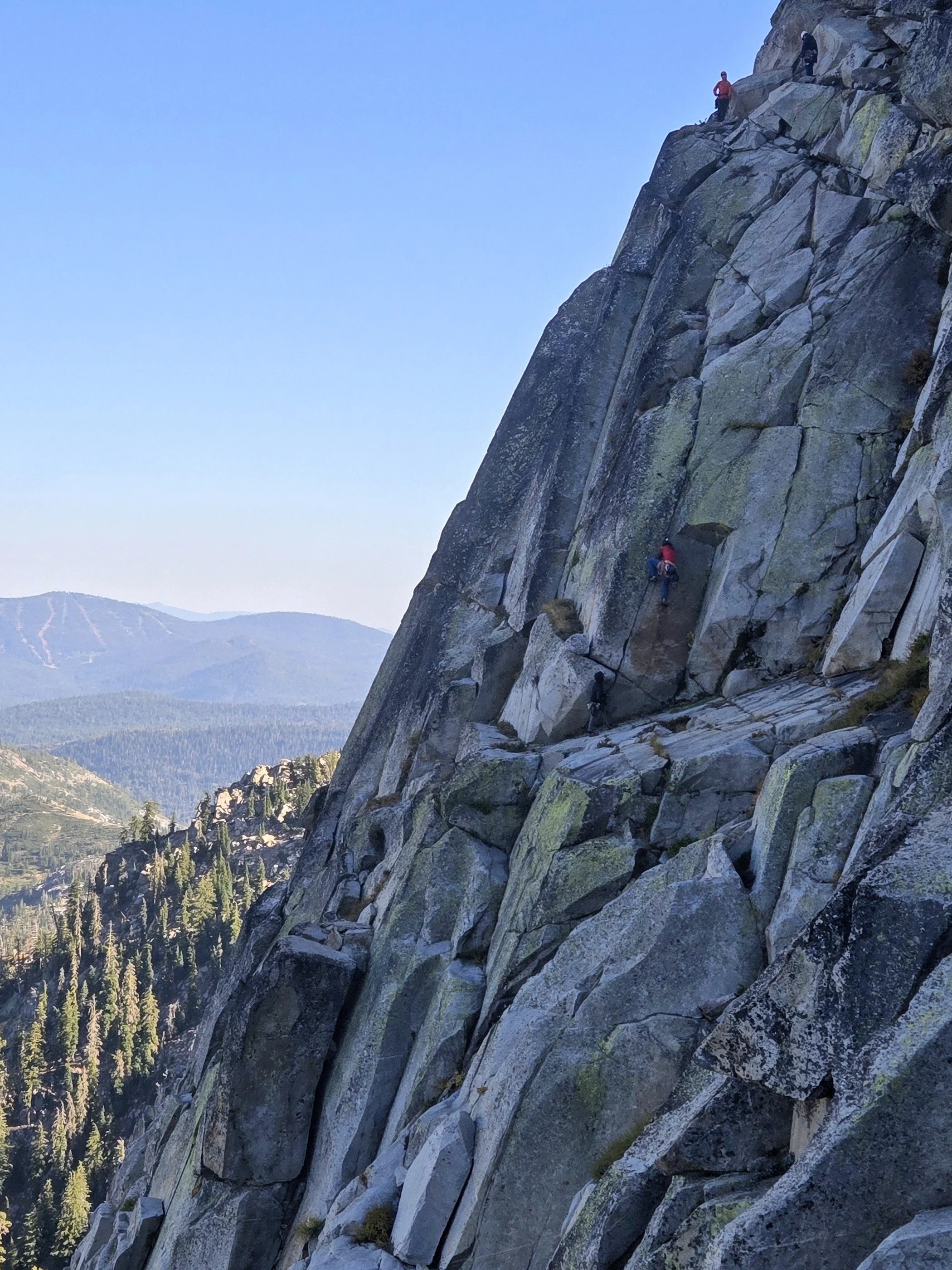 People rock climbing on a steep granite cliff with a mountain landscape in the background.