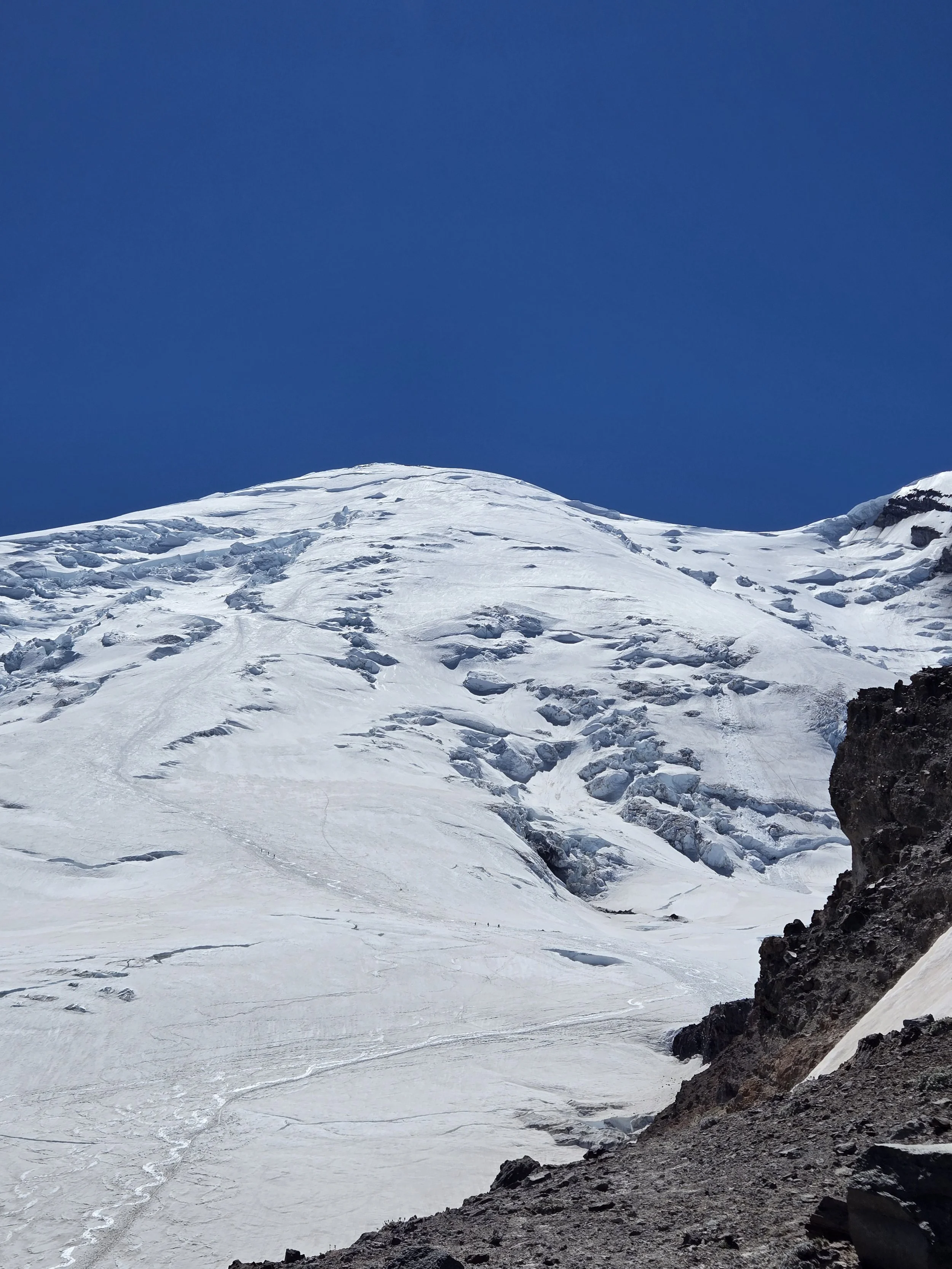 Snow-covered mountain peak under a clear blue sky with dark rocky terrain in the foreground.