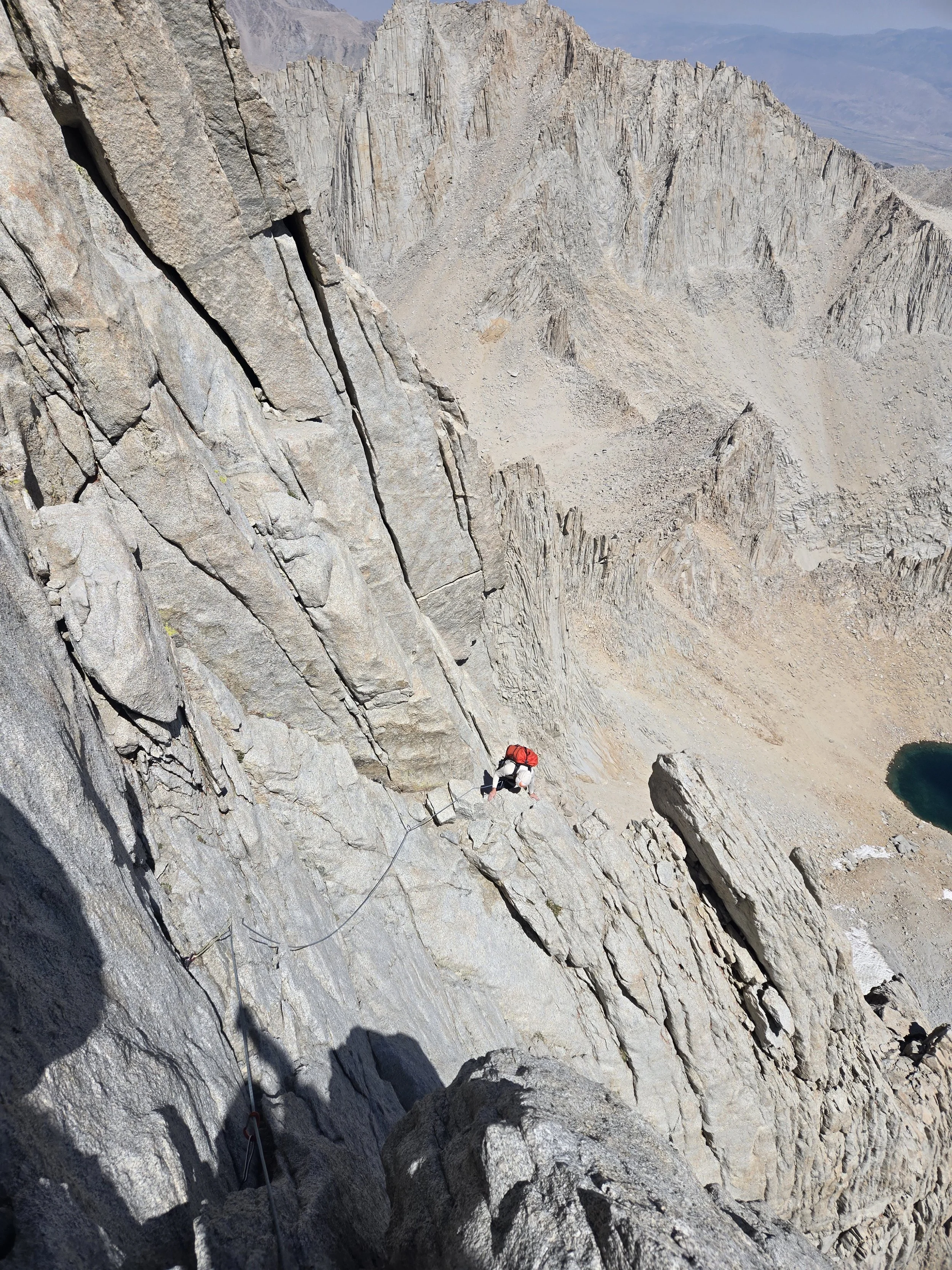 A climber wearing a red helmet and backpack ascending a steep rocky mountain face with safety ropes, overlooking mountain valleys and a small teal lake in a remote wilderness.