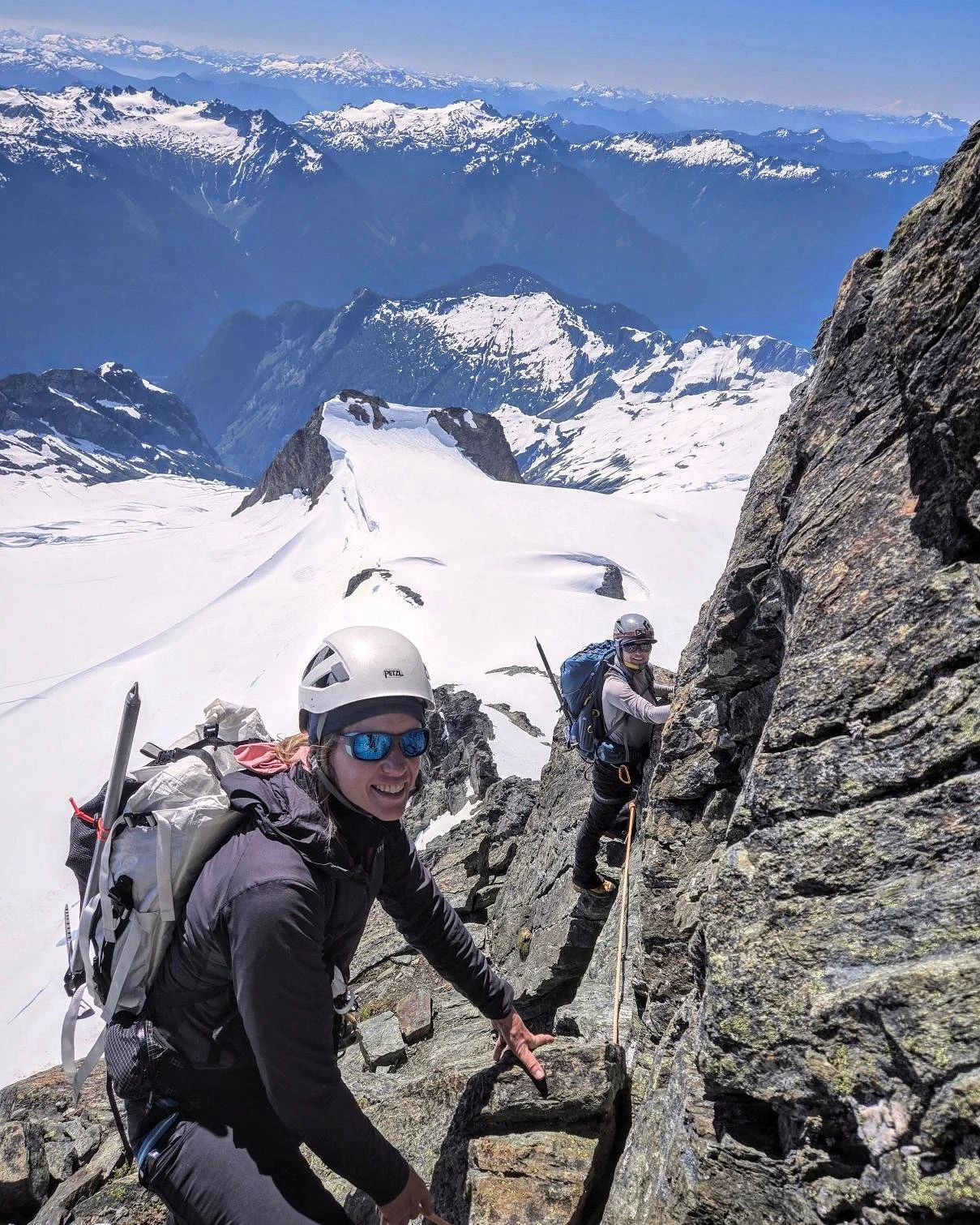 Two climbers ascending a rocky mountain face on a snowy peak, with a mountain range and snow-covered peaks in the background.