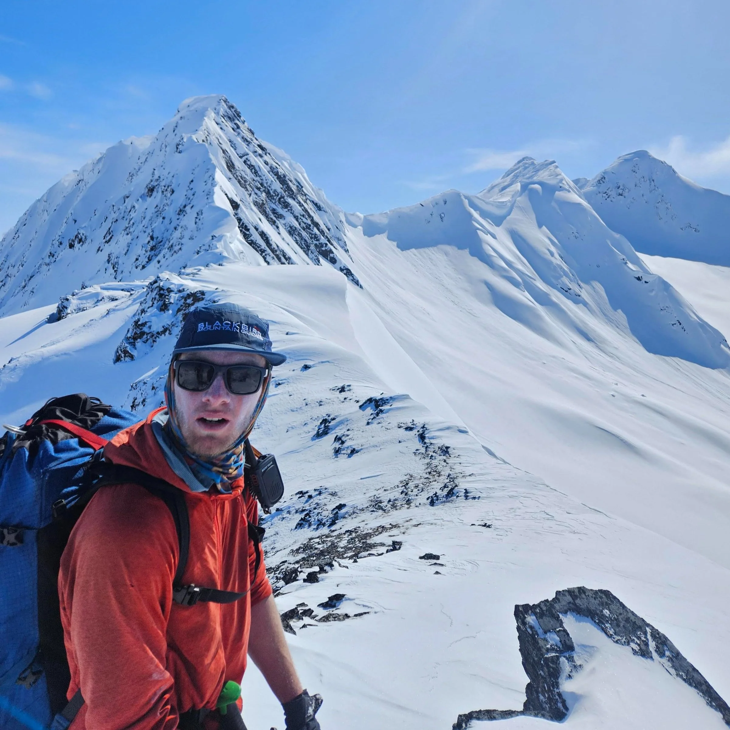 A man wearing sunglasses and outdoor gear stands on a snowy mountain ridge with snow-capped peaks in the background during daytime.