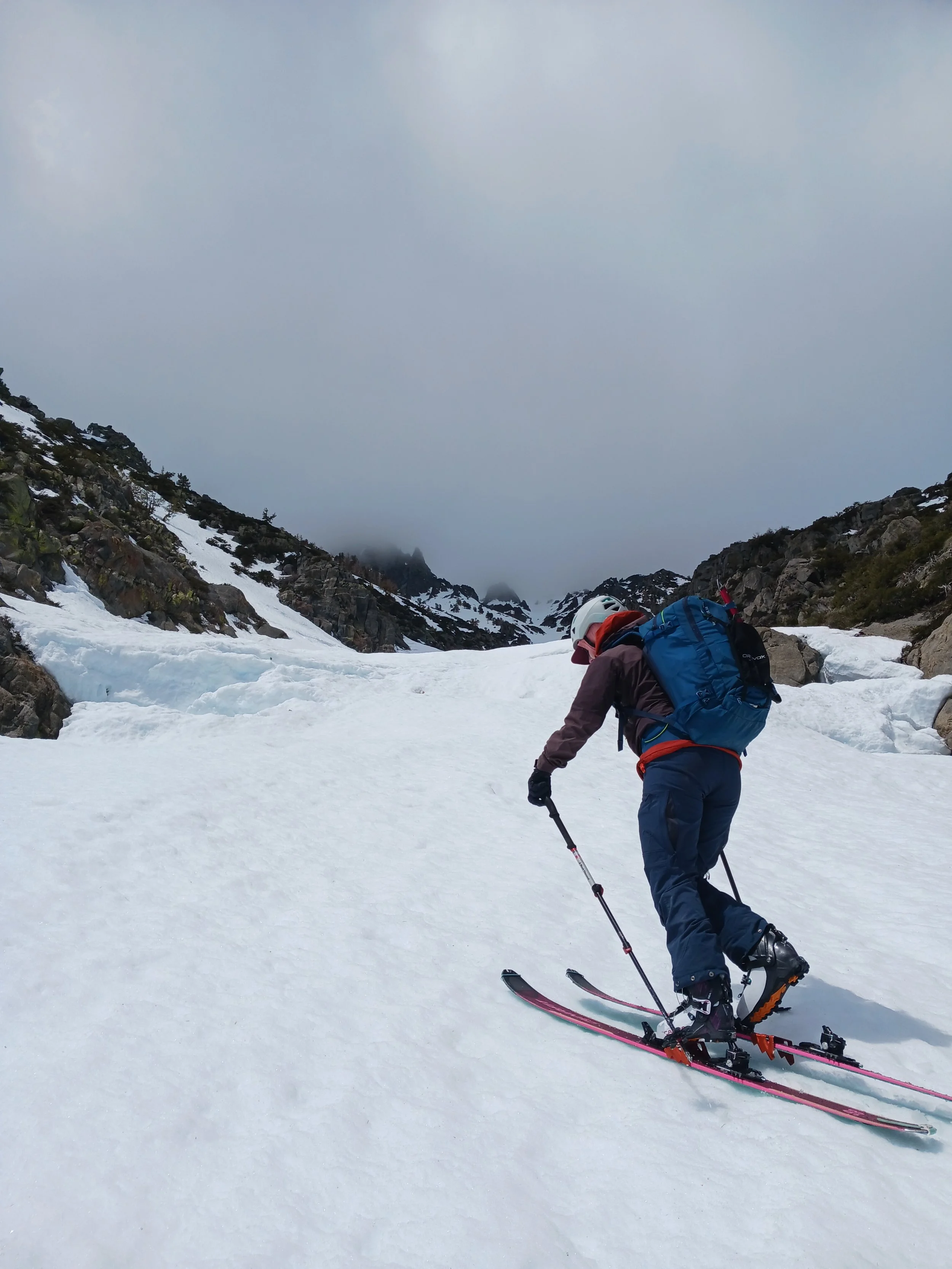 A person skiing on a snowy mountain slope with rocky terrain on either side, wearing a white helmet, dark brown jacket, blue pants, and carrying a blue backpack.