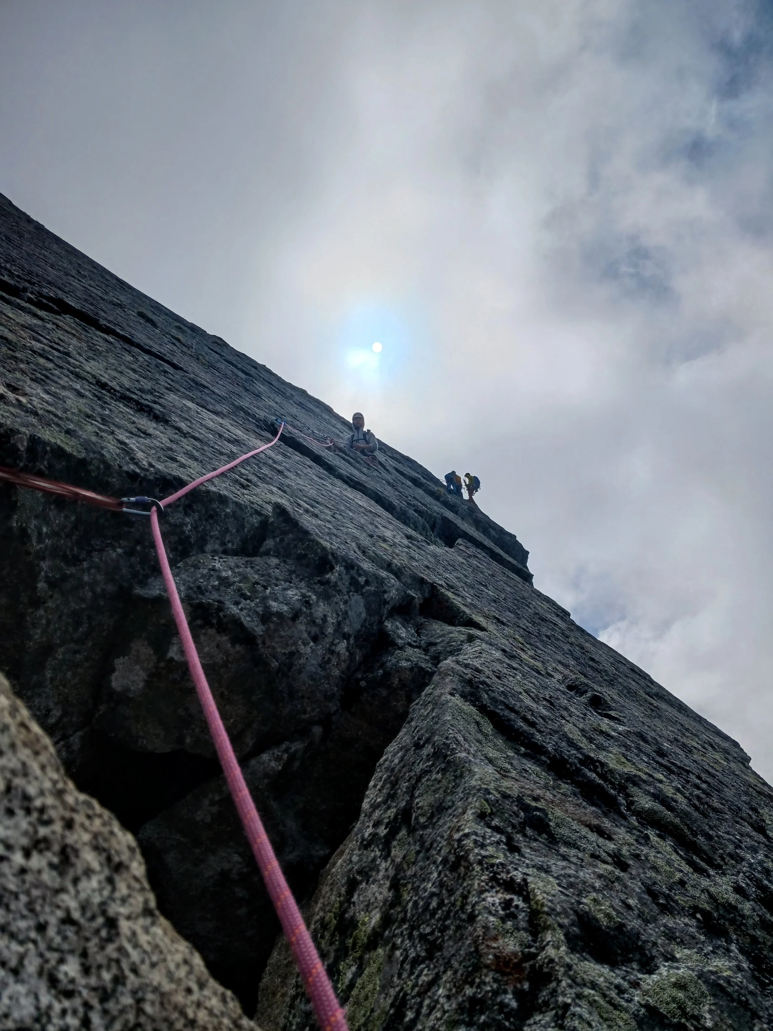 Rock climber ascending a steep cliff with a safety rope, viewed from below with the sky and sun in the background.