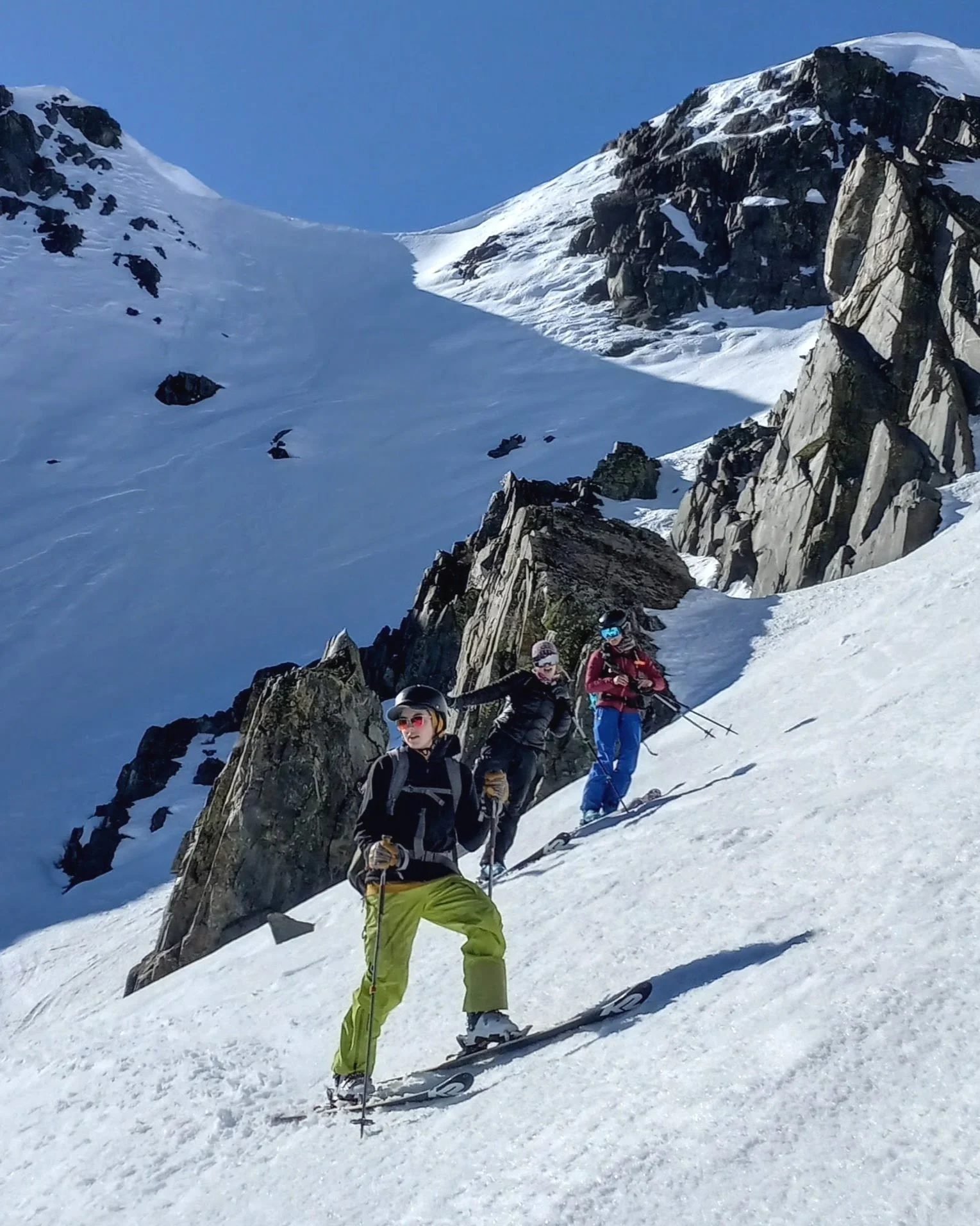 Three people skiing on a snowy mountain slope with rocky cliffs and snow-covered peaks in the background.