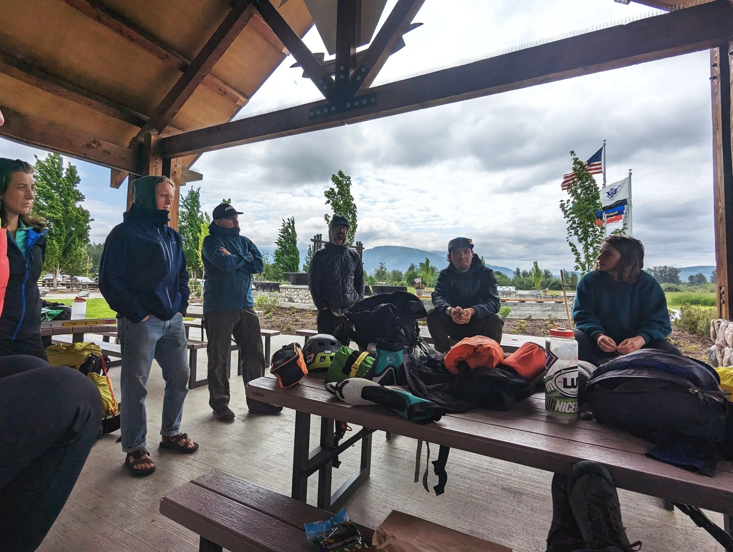 Group of six people gathered under a wooden shelter, with some sitting and others standing, outdoors on a cloudy day with flags in the background. The table has gear including helmets, backpacks, and water bottles.