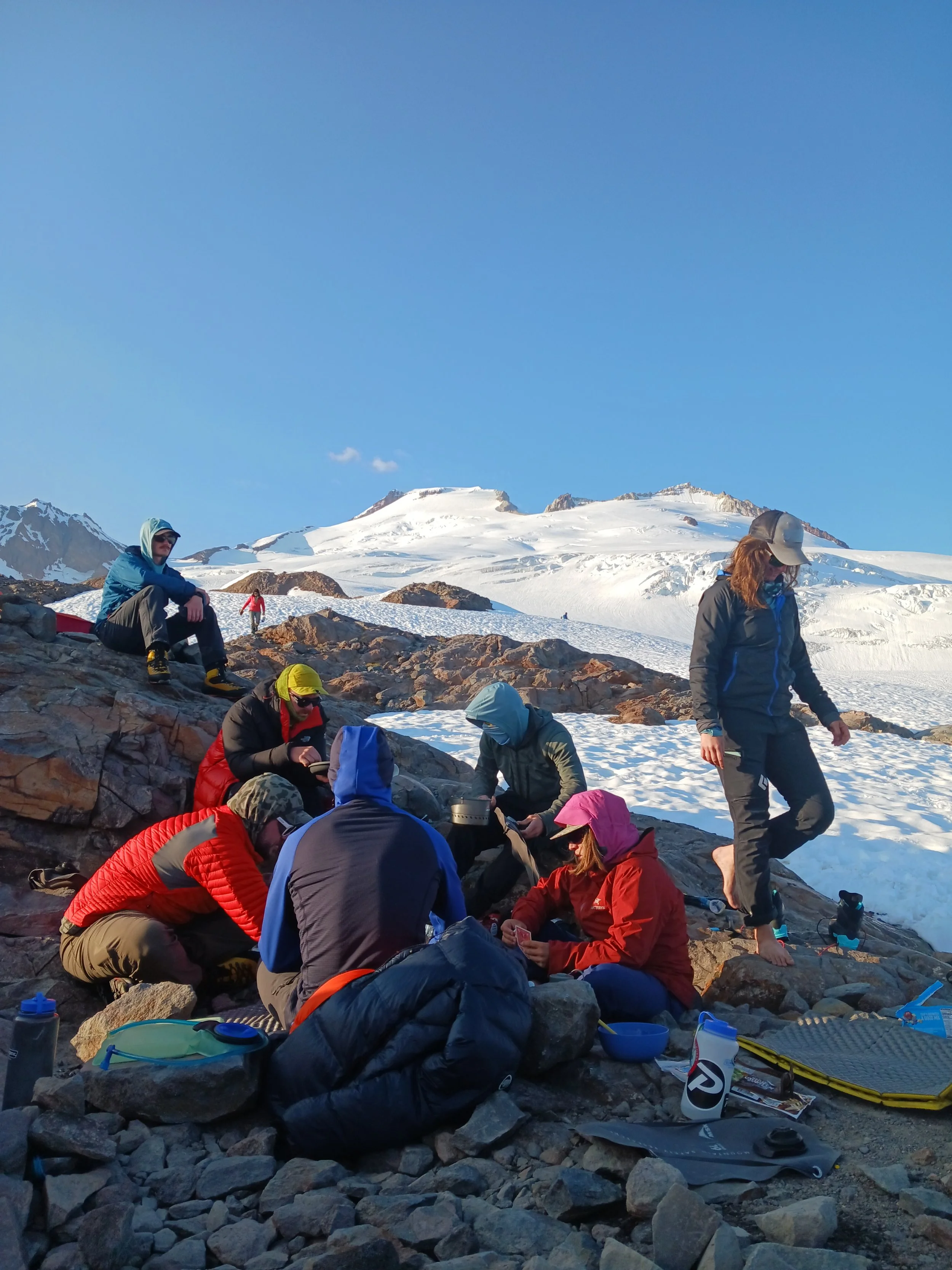 A group of hikers resting on rocky terrain surrounded by snow with snowy mountains in the background on a clear day.