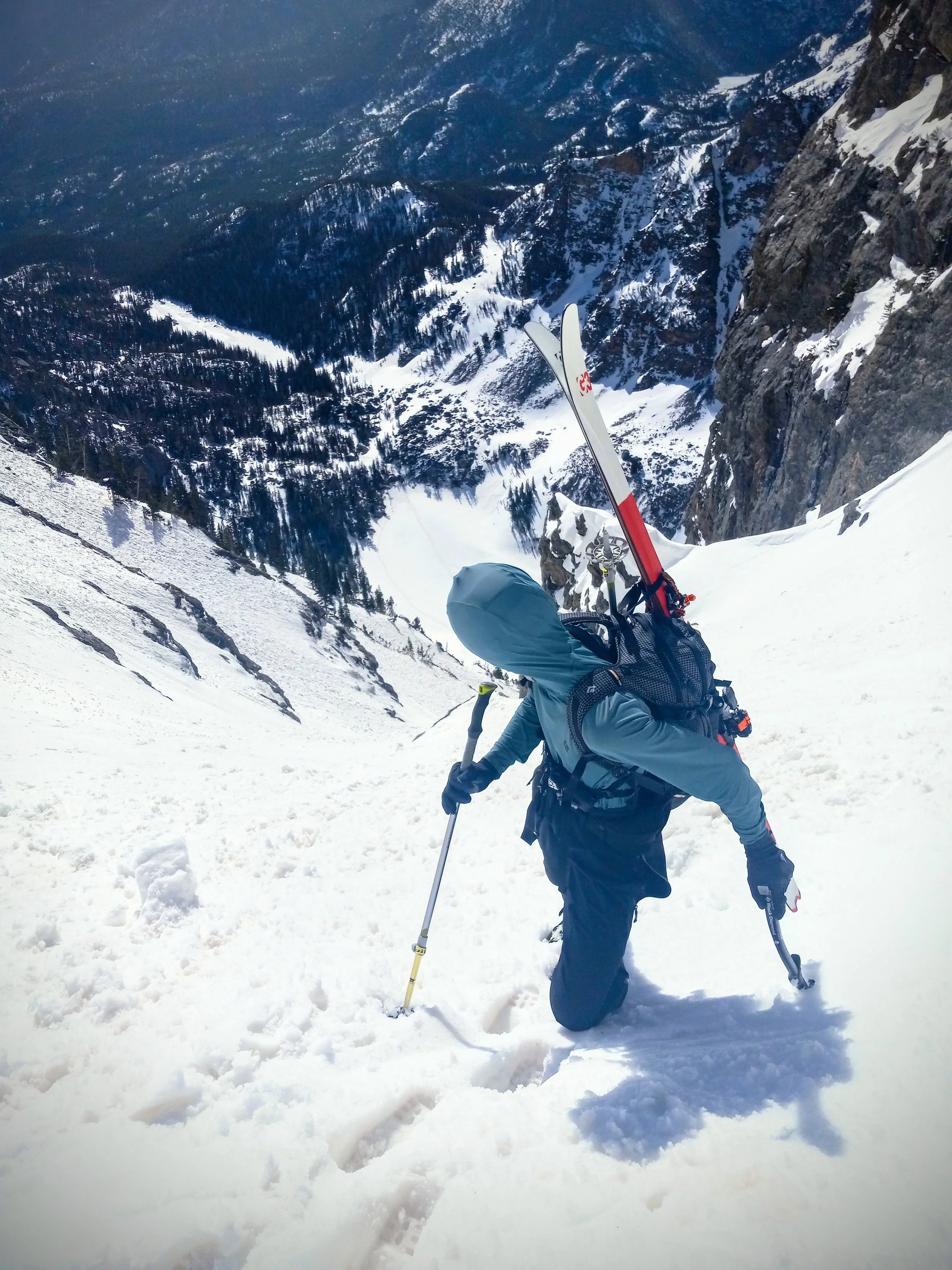 A climber in outdoor gear ascending a snowy steep mountain slope with skis on their backpack, overlooking a snowy valley and forested mountains.