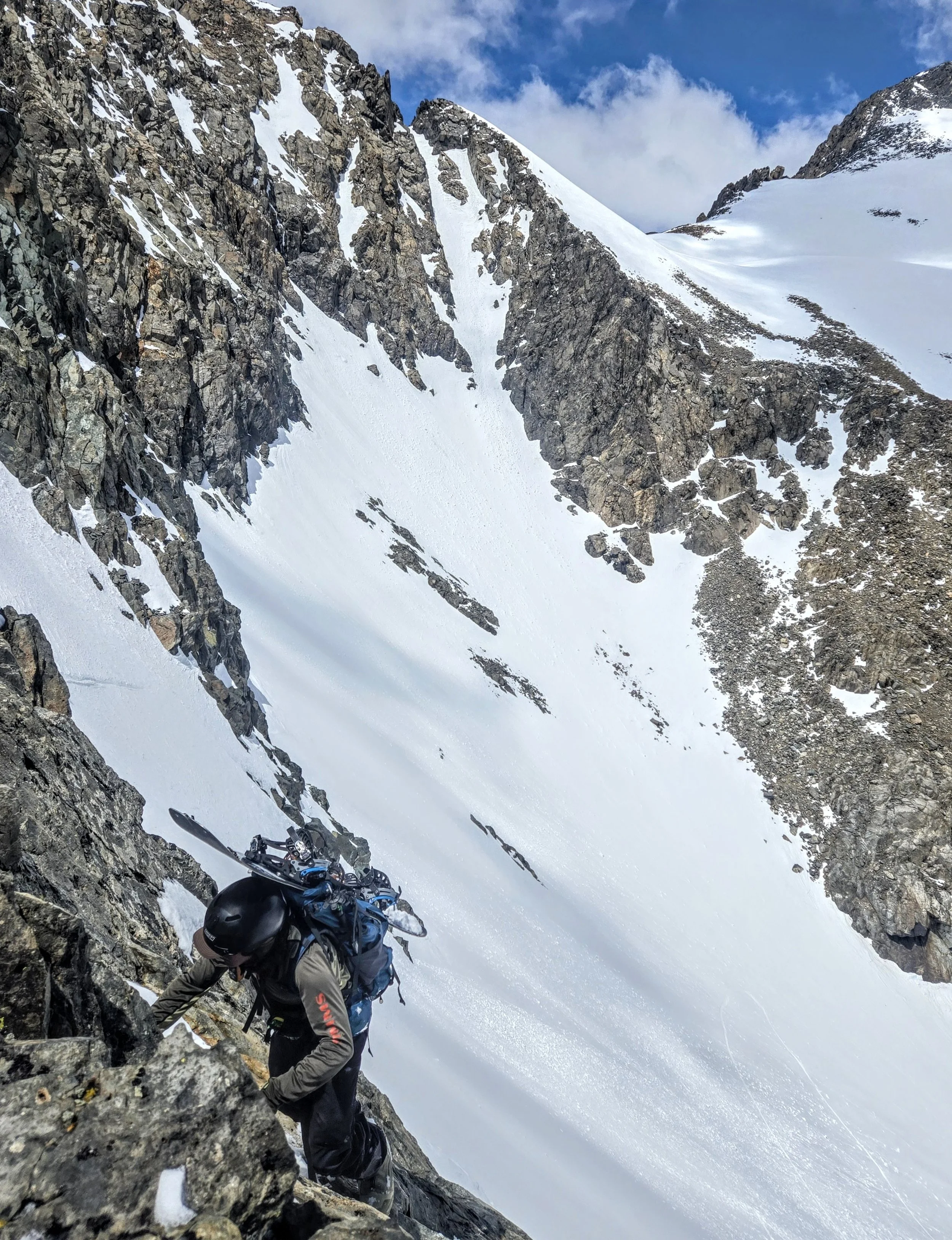 A climber ascending a rocky mountain with snow, carrying gear, with a snow-covered slope and rugged cliffs in the background.