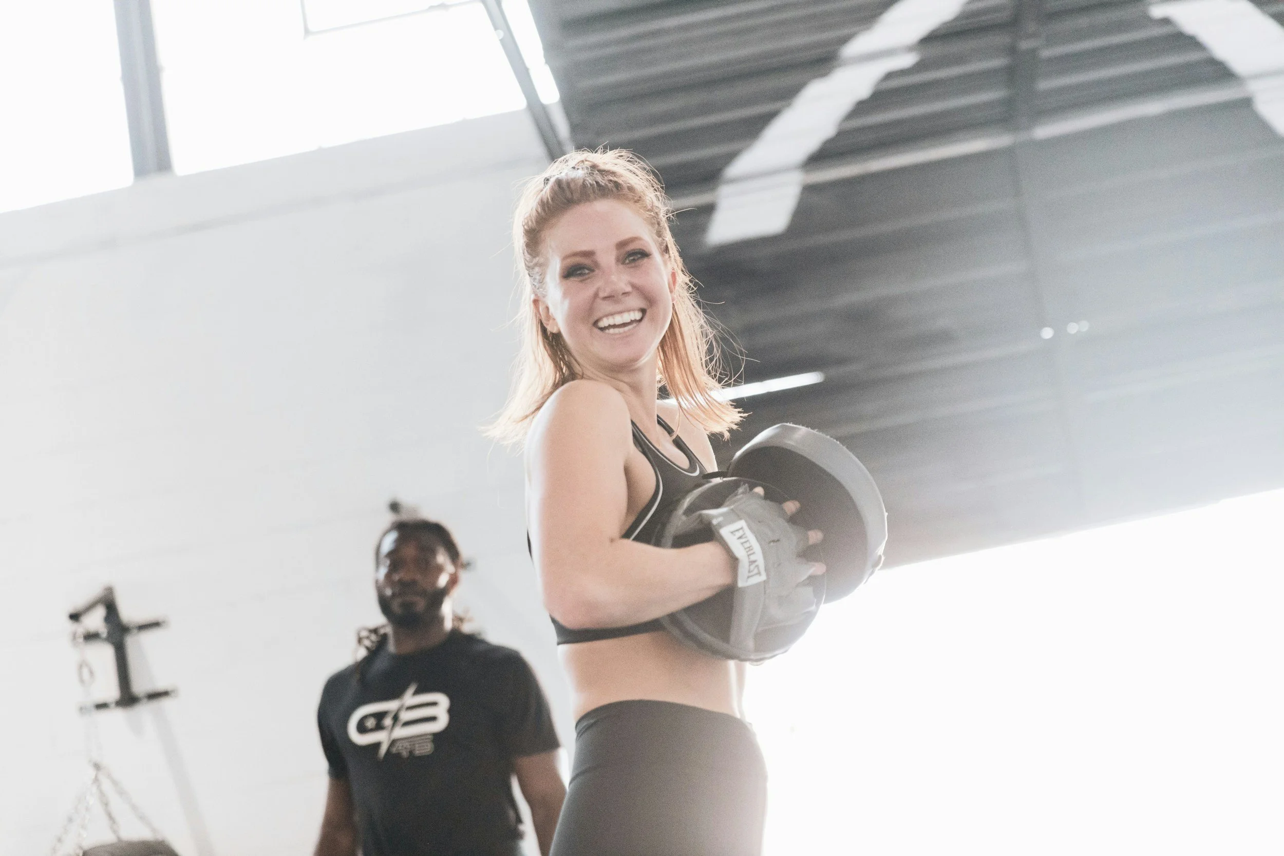 A happy woman holding a weight plate in a gym, with a trainer standing behind her.