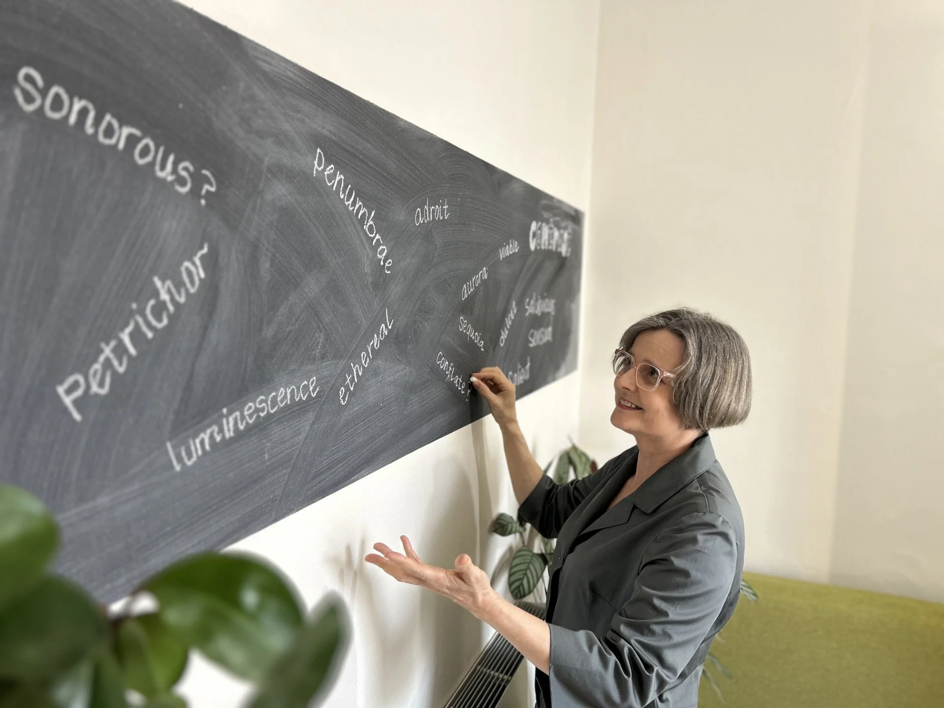A woman with short gray hair and glasses writing on a black chalkboard in a room with white walls and green plants.