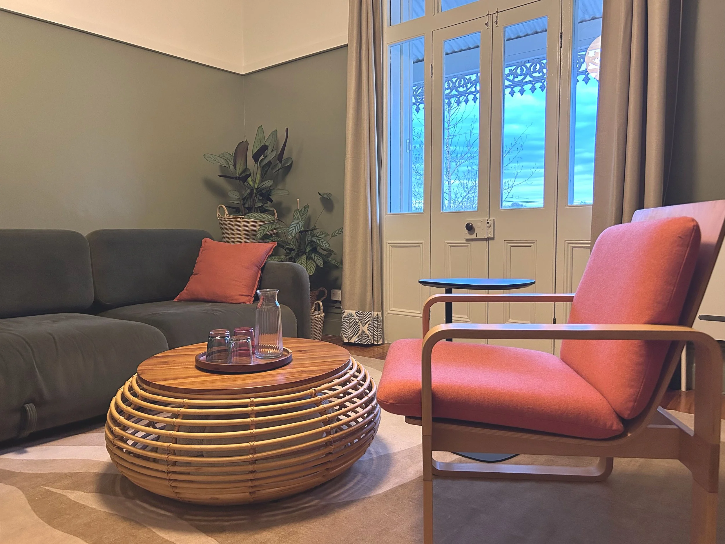 Living room with a dark green sofa, orange cushion, round wooden coffee table with glass jars, potted plant in a basket, and pink armchair. Large window with curtains reveals a cloudy sky.
