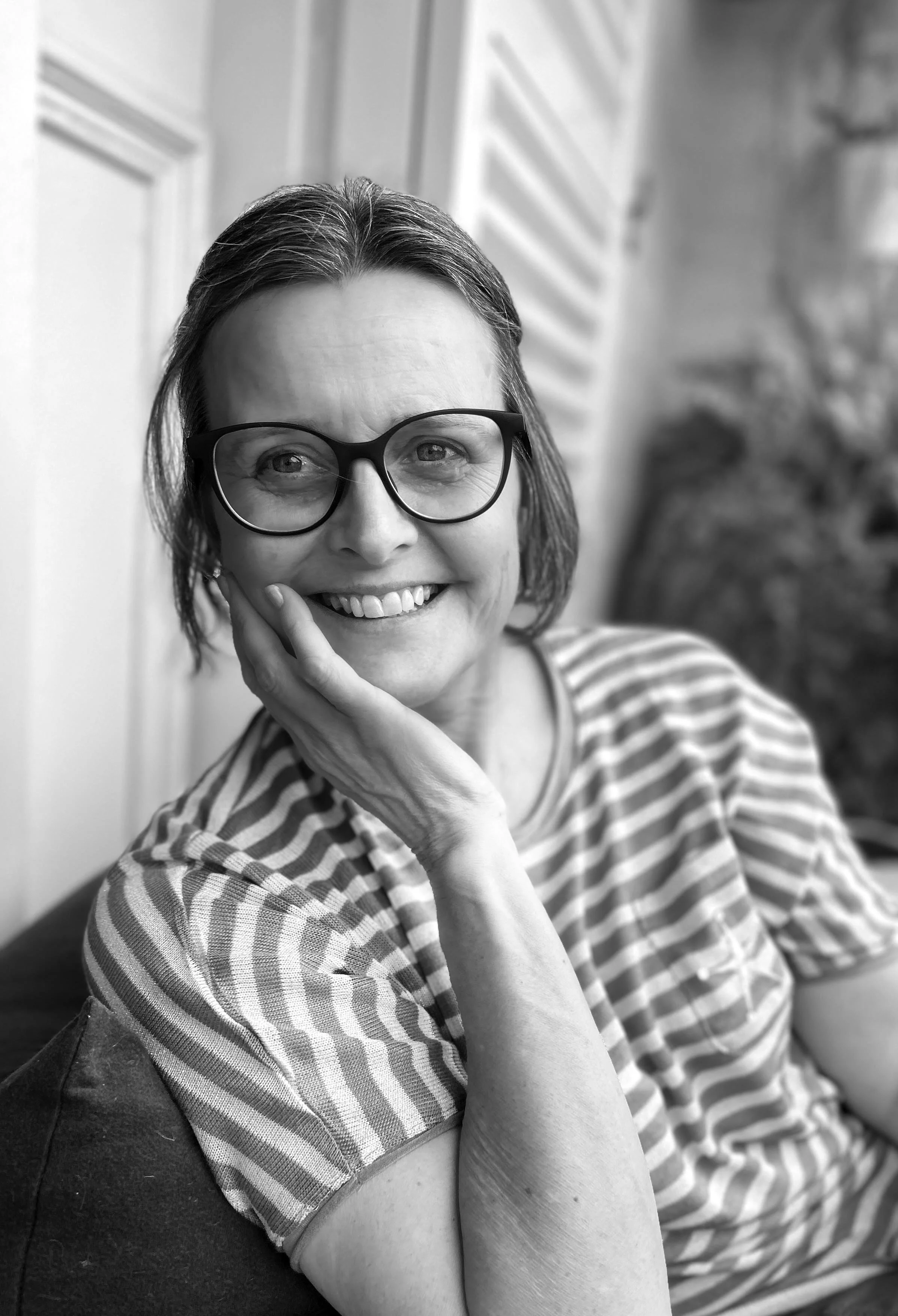 A black and white photo of a smiling woman with glasses, resting her chin on her hand, wearing a striped shirt.