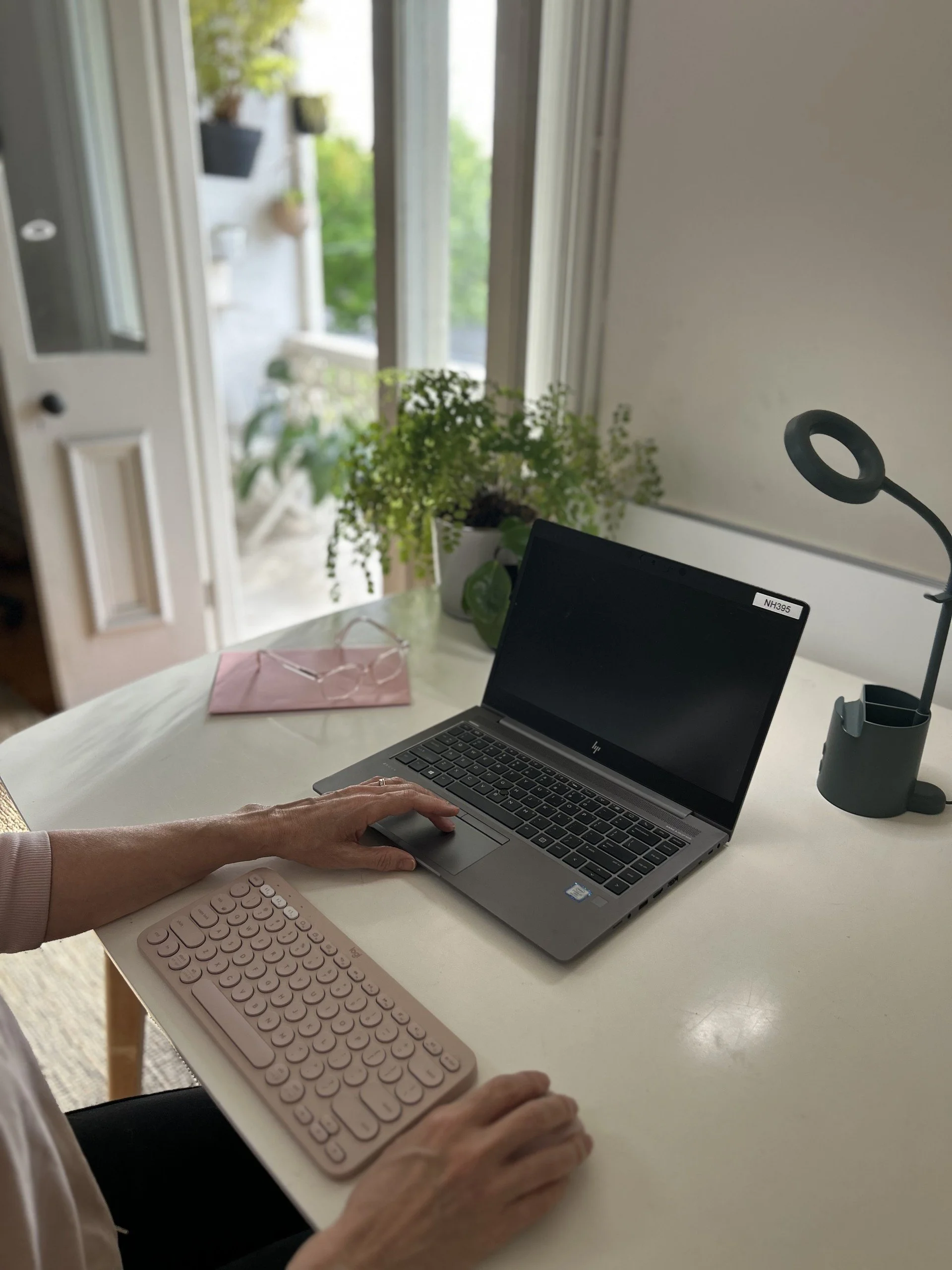 A person using a laptop at a white table with a pink wireless keyboard and a pink mouse, near a window and a potted plant.