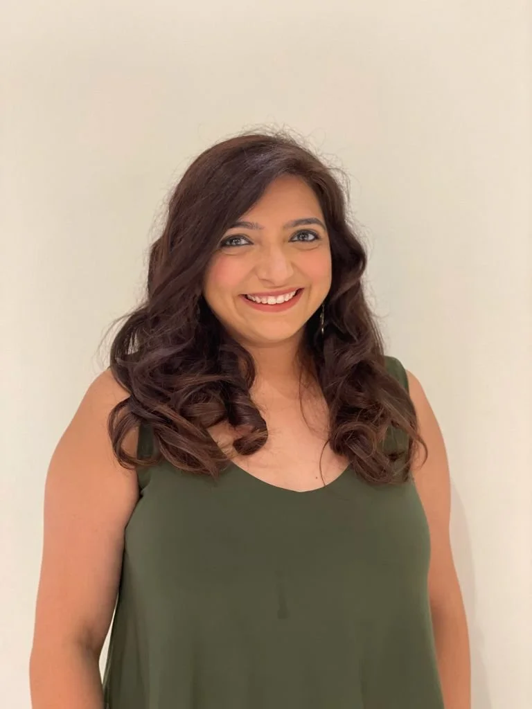 A woman with long, curly dark hair and light skin, smiling, wearing a sleeveless green top, standing against a plain light-colored background.