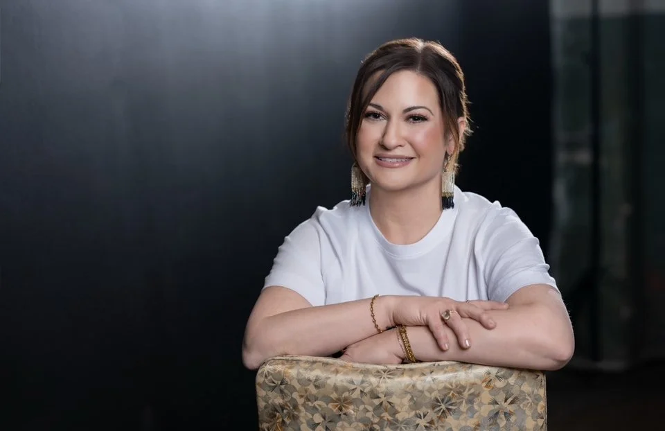 A woman with short brown hair and earrings sitting at a table against a dark background, smiling and looking at the camera.