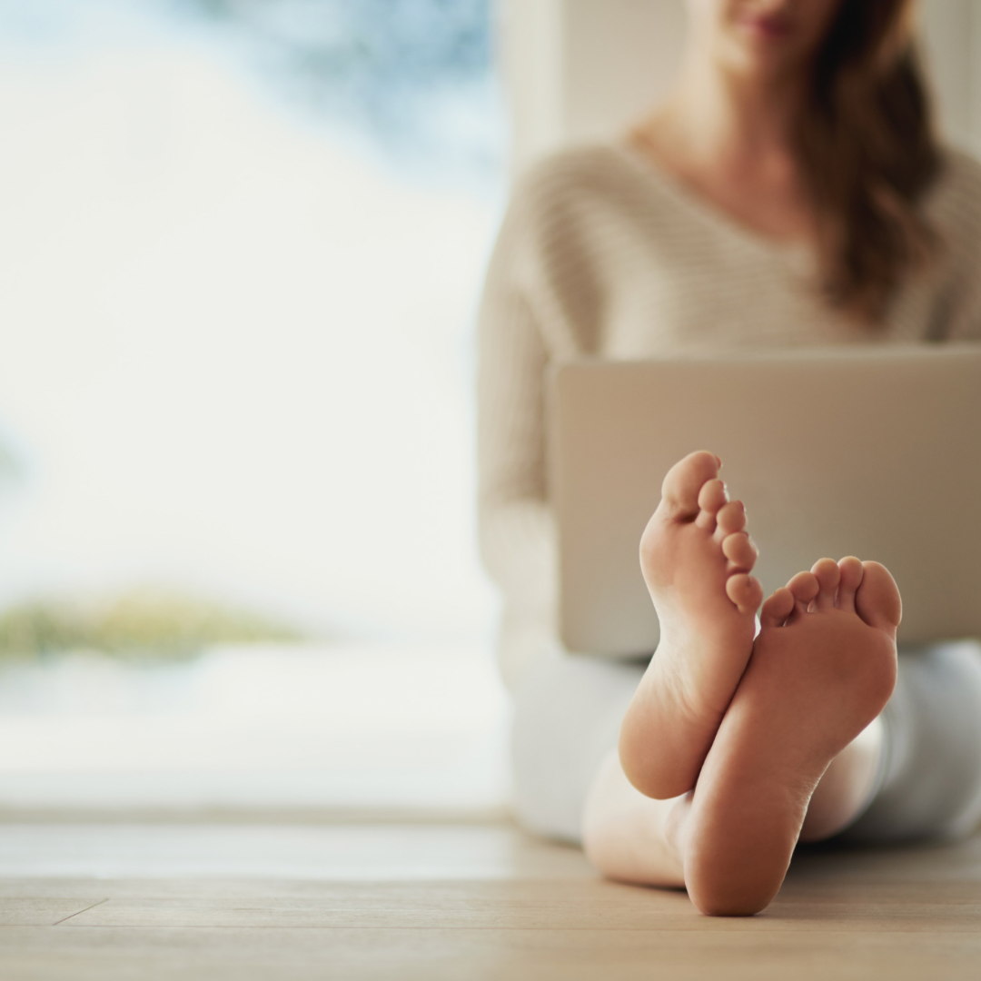 Person sitting on the floor using a laptop with their feet extended towards the camera, in a bright room.