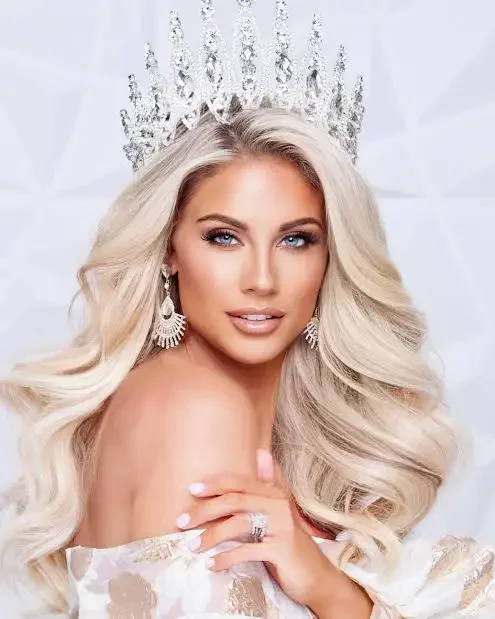 A woman with long blonde hair, wearing a large crystal crown, diamond earrings, and an off-the-shoulder dress, posing with her hand on her chest against a white background.