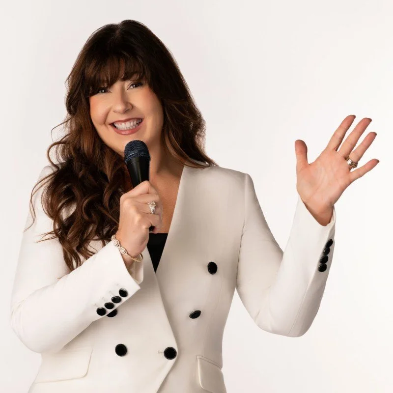 A woman with long brown hair holding a microphone and smiling, dressed in a white blazer, waving with her other hand.