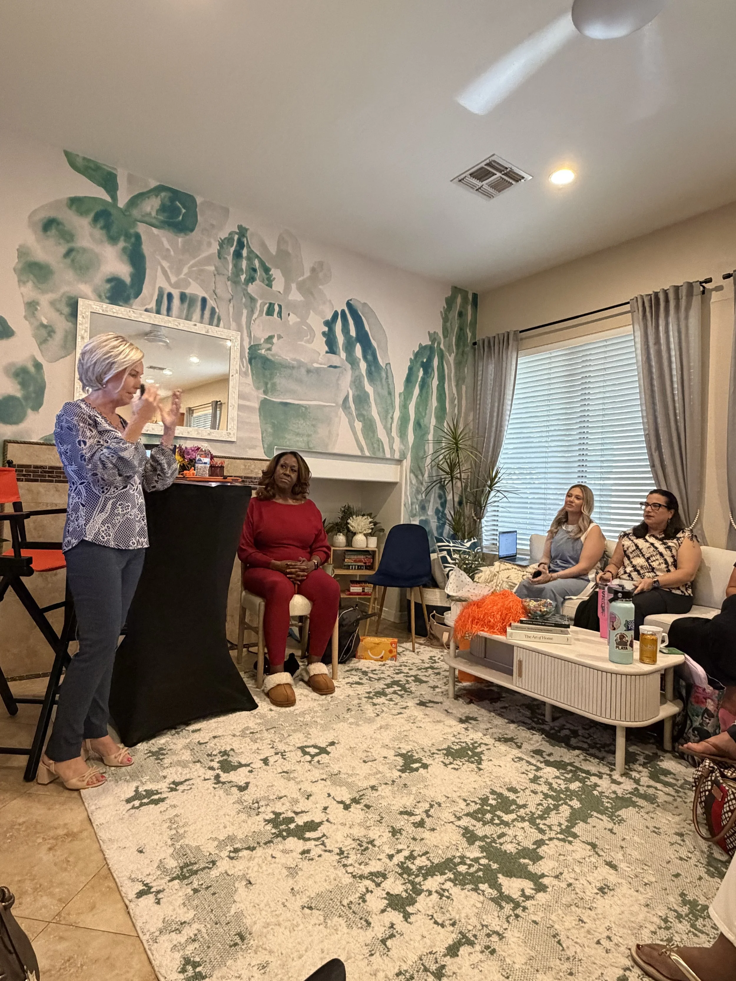 A group of women sitting and standing in a living room with a painted wall mural of potted plants, a large window with curtains, and various objects on a coffee table and nearby surfaces. One woman appears to be speaking while others listen.