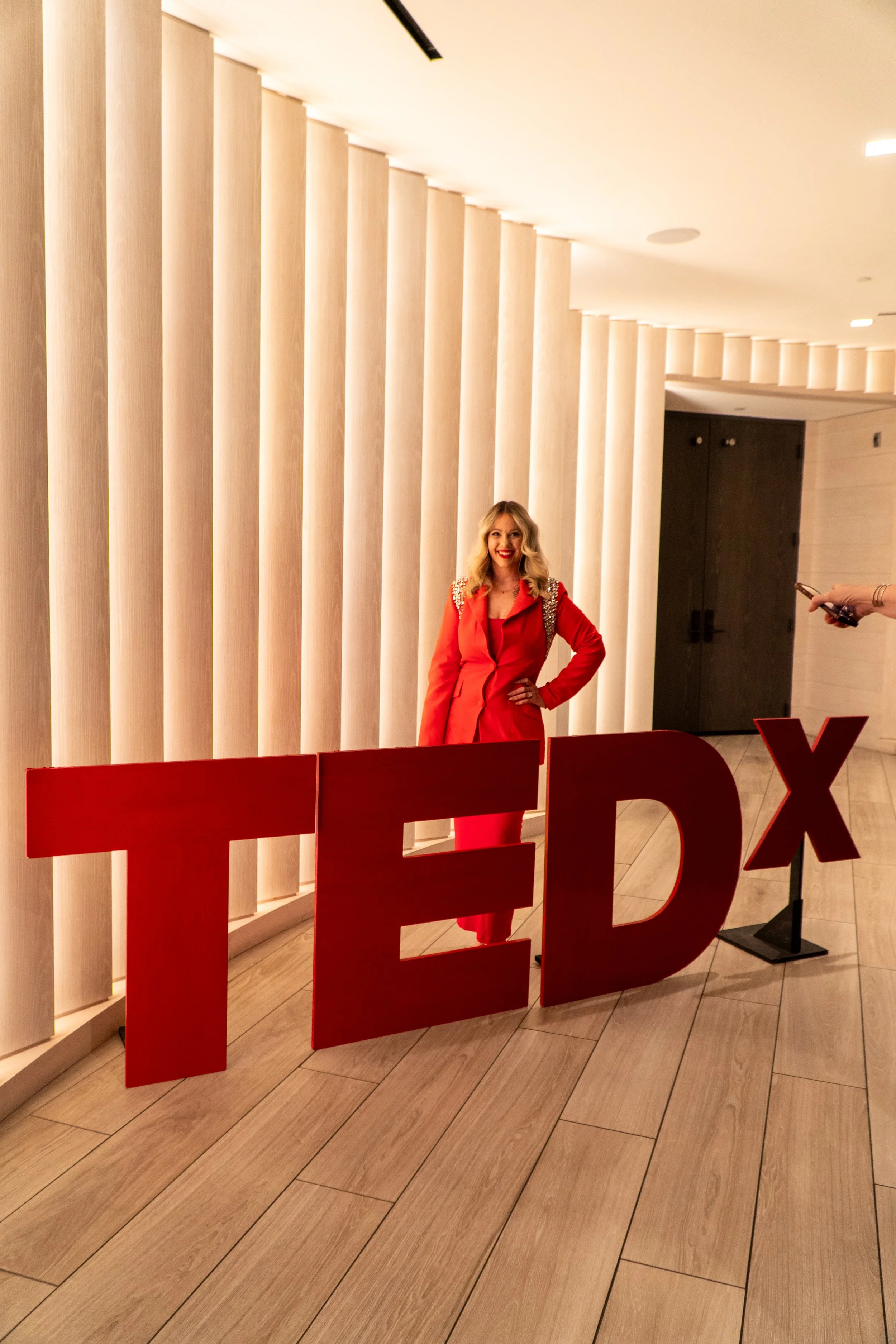 A woman in a red outfit is standing behind large red TEDx letters in an indoor venue with warm lighting and a modern design.