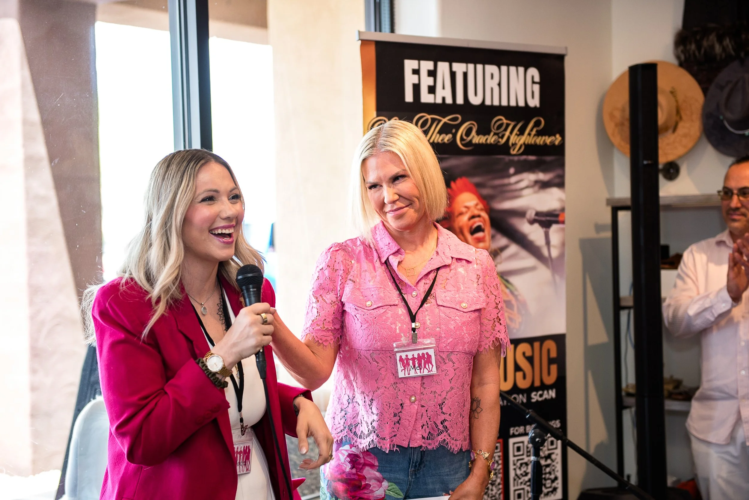 Two women are standing close together and smiling; one woman is wearing a pink lace top and the other is in a red blazer. They are at an event with a black sign behind them that says, 'FEATURING Thie Oedes Nheiwaver' and a photo of a singer. One woman is holding a microphone.