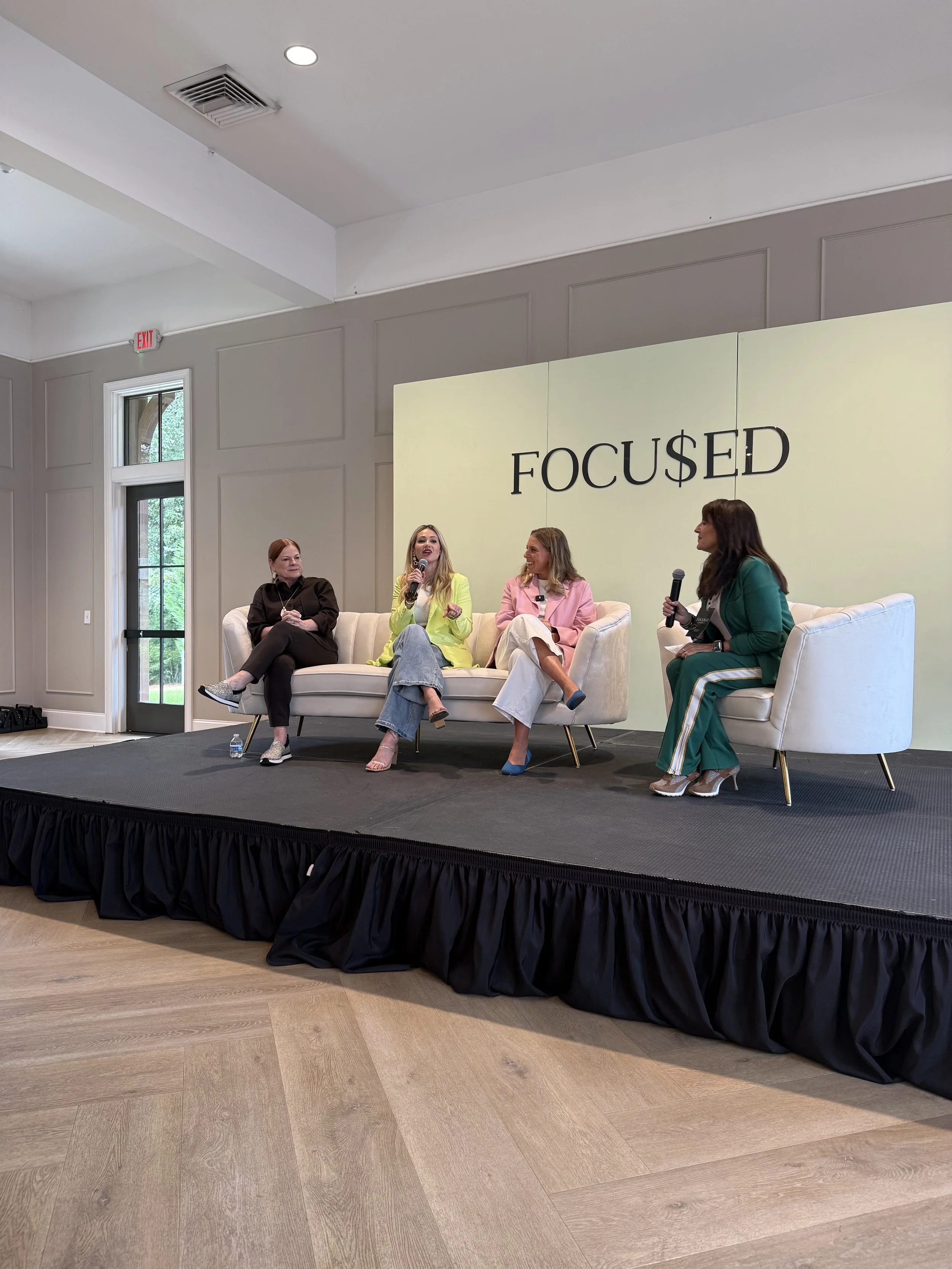 Four women are sitting on a stage with a large sign behind them that reads 'FOCUSED.' They are engaged in a discussion, with one woman speaking into a microphone. The stage has a black skirt, and the room has large windows and a wooden floor.