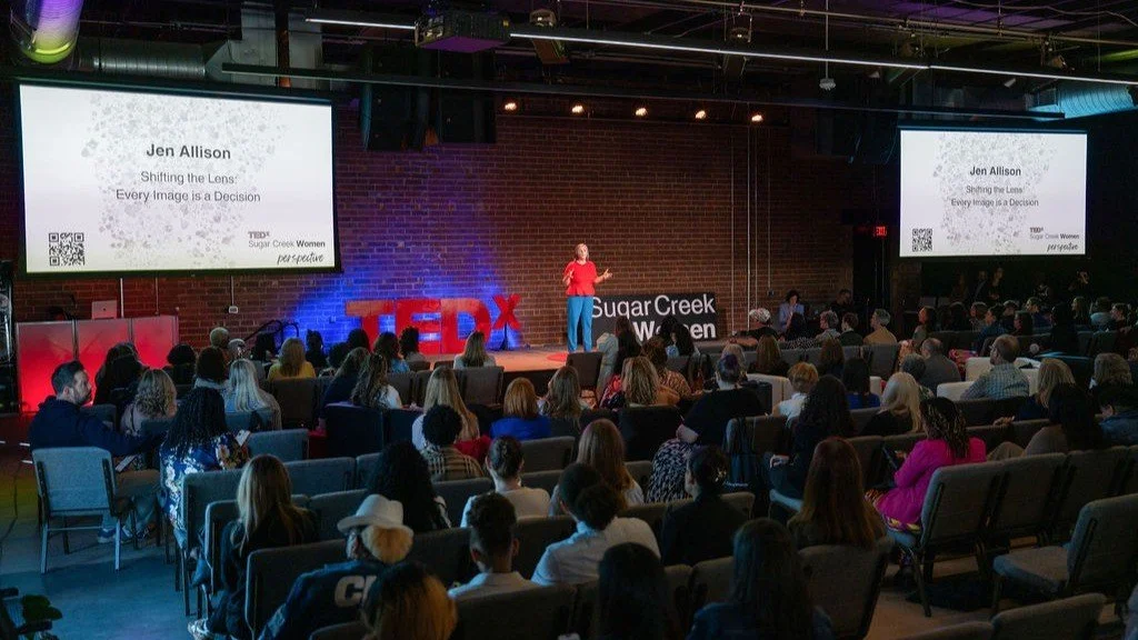 A woman is giving a TEDx talk in a large venue with a brick wall background and large screens displaying her name, Jen Allison, and title, 'Shifting the Lens: Every Image is a Decision.' An audience sits facing the stage, and large red TEDx letters are on stage behind her.