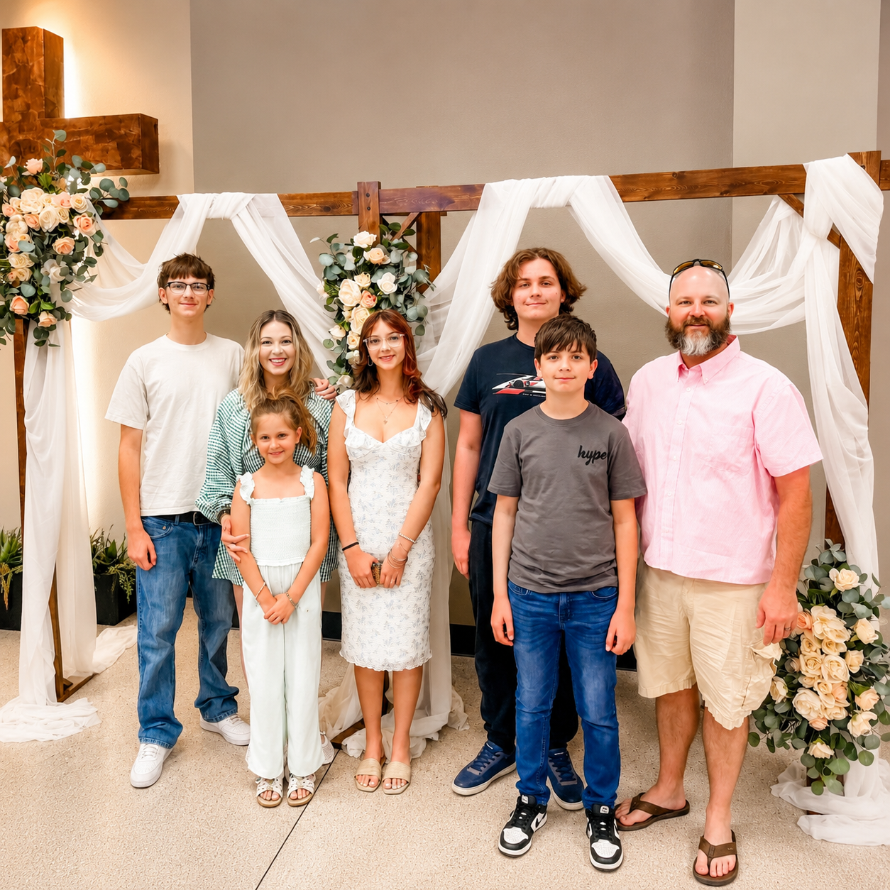 A group of seven people standing in front of a floral wedding arch with white drapery, including four children and three adults, outdoors, at a celebration or wedding event.