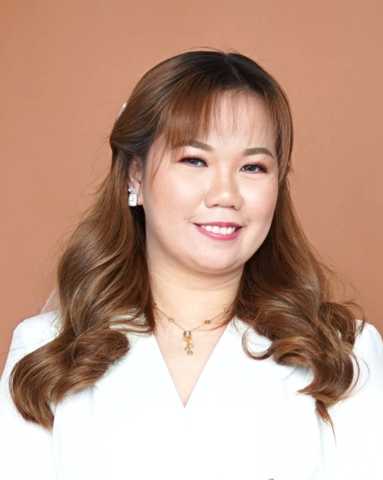 A woman with brown hair styled in loose waves, wearing a white top, earrings, and a gold necklace, smiling against a plain background.