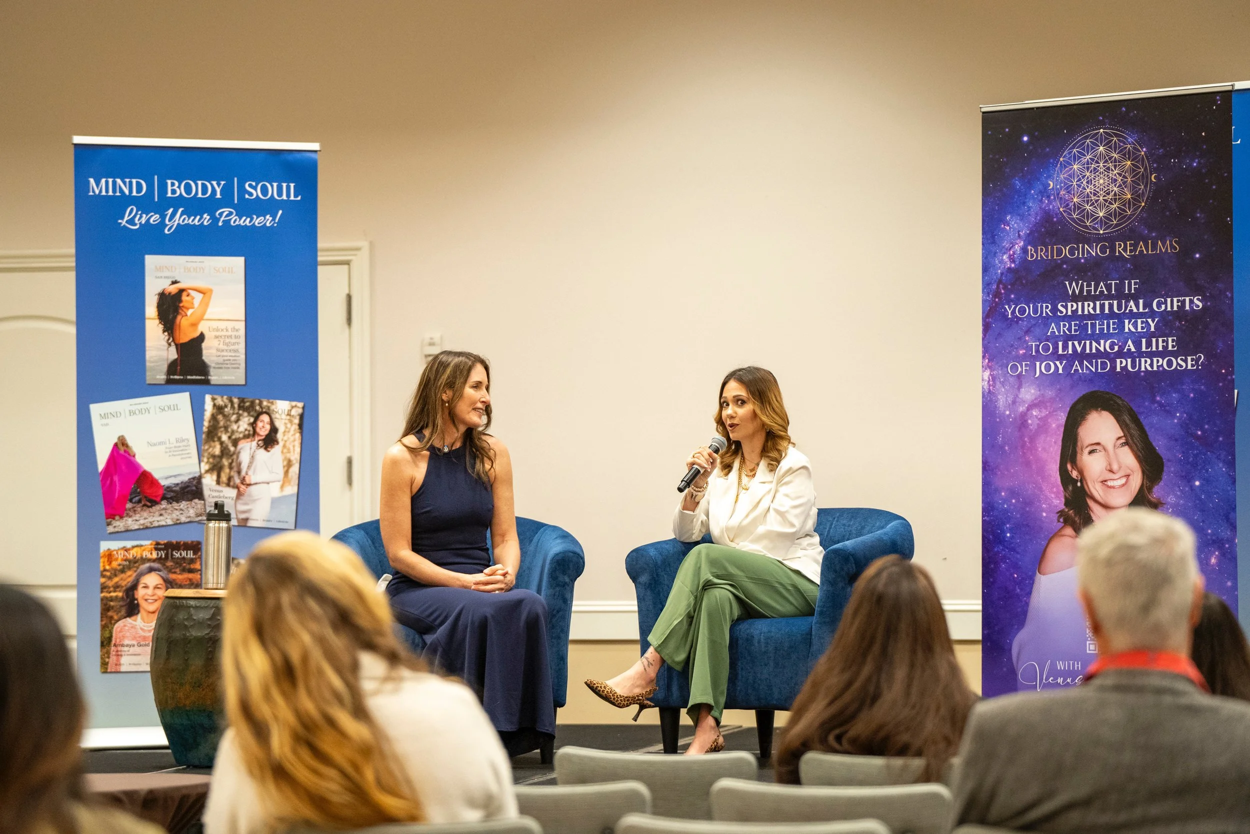 Two women seated on stage engaged in conversation during a panel discussion at an event. The woman on the left has long brown hair and is wearing a navy blue dress, while the woman on the right has long light brown hair and is wearing a white blazer and green pants, holding a microphone. Behind them are two large vertical banners with promotional content, one blue and one galaxy-themed, and a small round table with a water bottle in front of the woman on the left. Audience members are visible in the foreground.