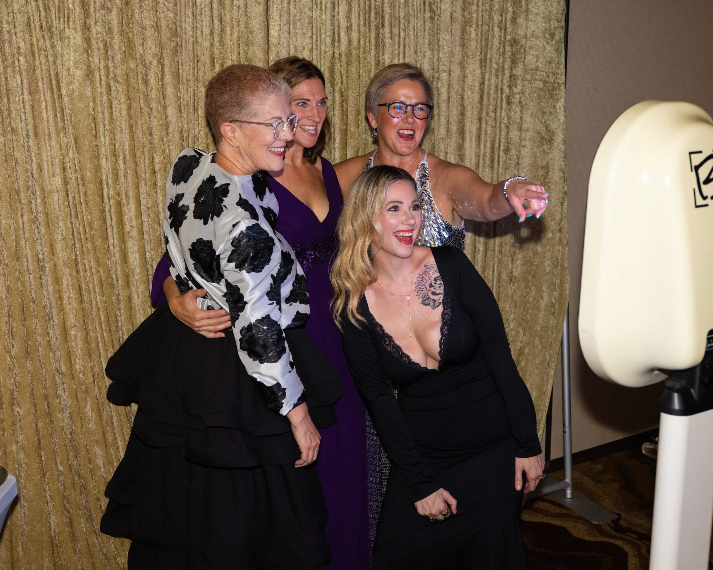 Four women are taking a group photo at an indoor event. They are smiling and posing in front of gold curtains. One woman is holding a large white balloon or prop with black writing on it.